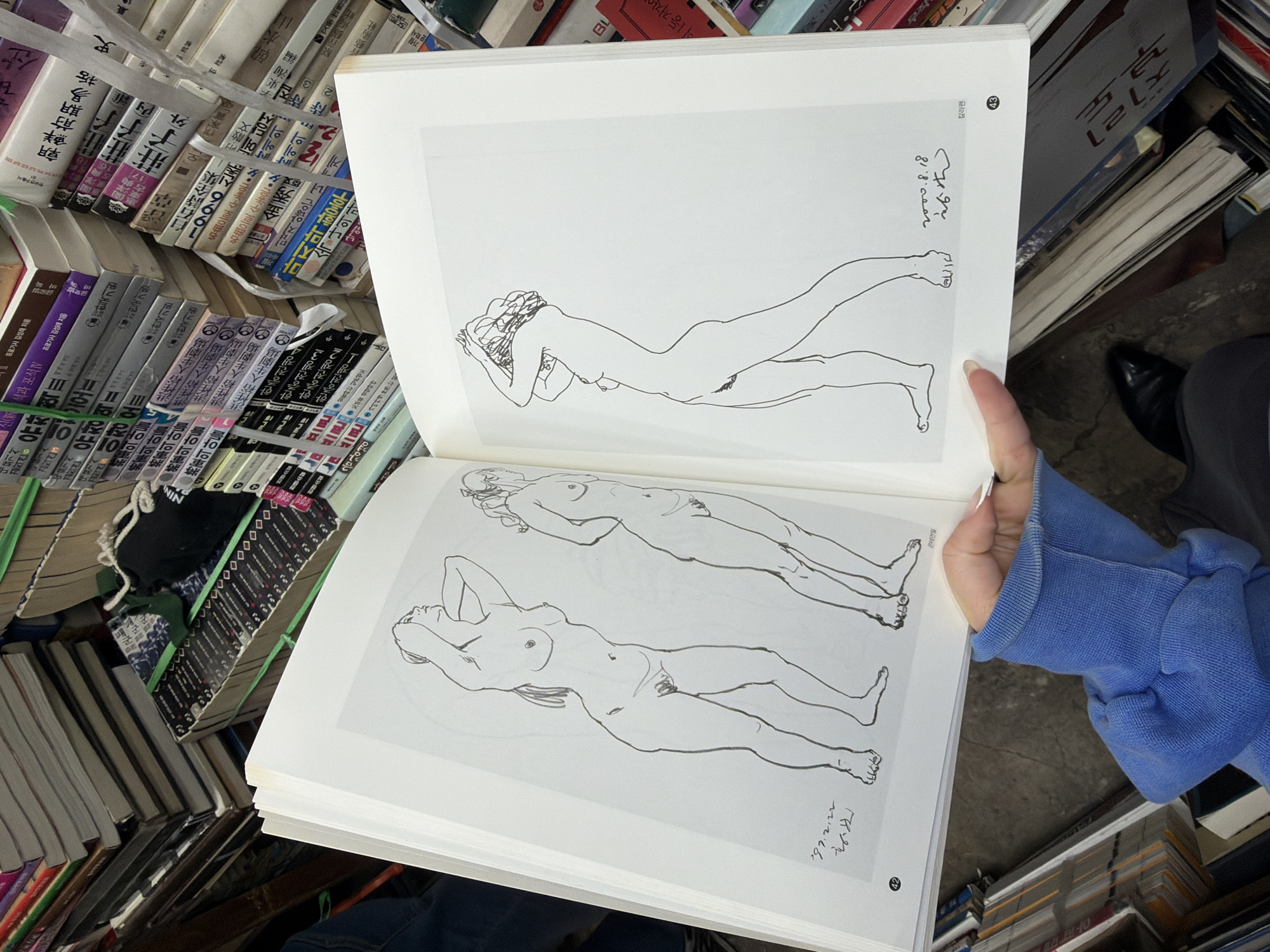The image captures a scene of a book being held open to display artwork, situated amidst stacks of books, likely in a bookstore or a second-hand book market. The book's pages show line drawings of nude figures in reclining poses. Korean text and numbers ("42", "43") are visible on the edges of the book pages.
The book is surrounded by numerous other books with Korean text on their spines, some bundled with green plastic straps. The books appear to be a mix of literary works, philosophy, and potentially other genres. A person wearing a blue sleeve is holding the book open. Part of a shoe and pants are also visible.
Based on the Korean text and book titles, the location is likely in Seoul, South Korea, possibly a used bookstore or an outdoor book market that is a common sight in Seoul.