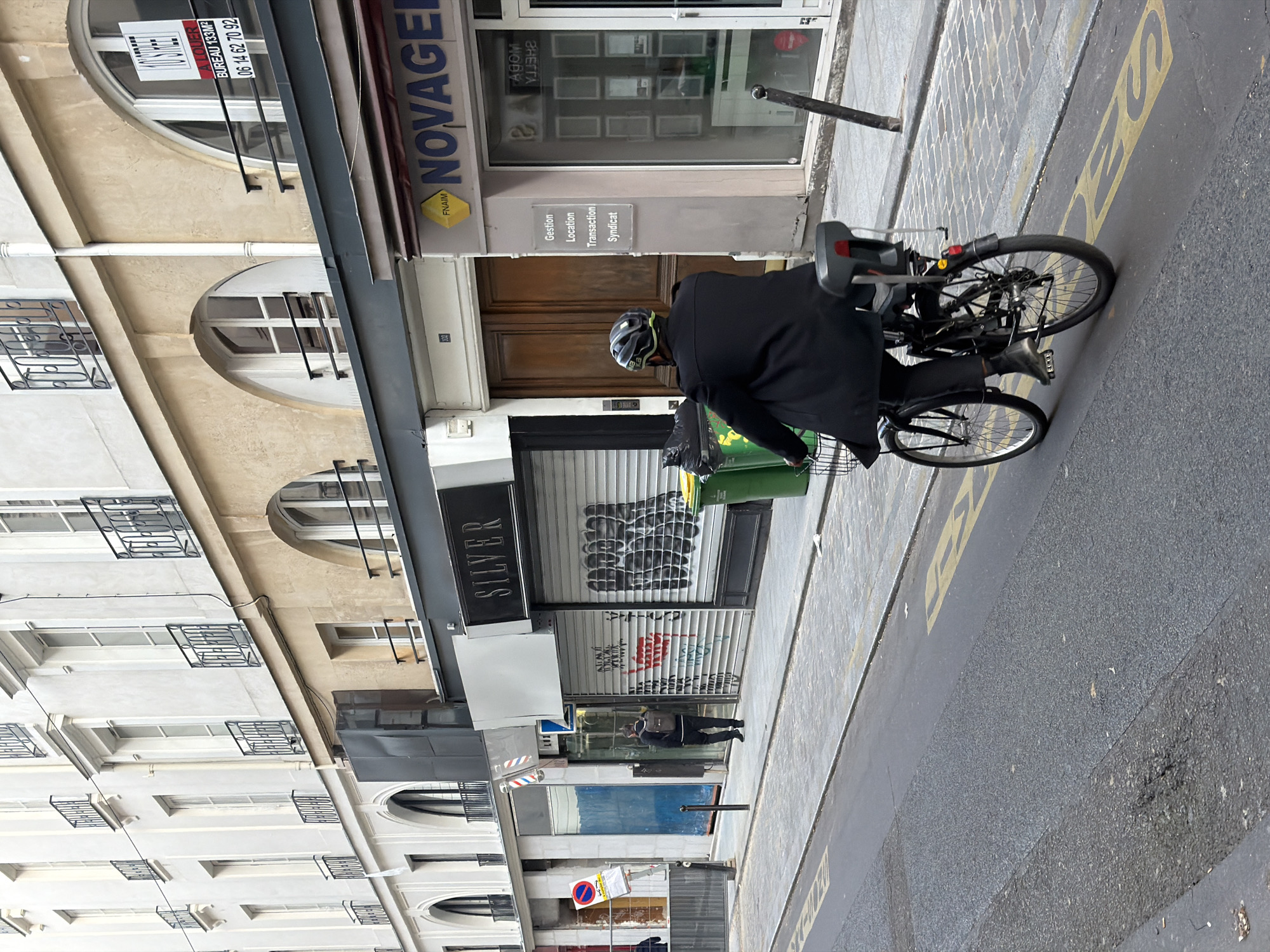 This street-level image captures a bustling urban scene, likely in Paris, France, characterized by its classic architecture and French signage. The perspective is slightly angled, showing a street lined with multi-story buildings housing businesses and residences.
In the immediate foreground, a person is on a black bicycle, wearing a dark coat and a grey and yellow helmet. The bicycle is equipped with a black child seat over the rear wheel and a basket or pannier area where a green trash bin and a black trash bag are visible. The cyclist appears to be either stationary or moving slowly along the curb. The road is asphalt with yellow painted markings, including partial letters such as "S" and possibly "DES".
The buildings on the left are typical Parisian Haussmannian style, featuring several floors with numerous windows, some arched, adorned with decorative iron grilles and balconies.
On the right side of the street, several commercial establishments are visible. A prominent business is "NOVAGEL," identified by its blue and yellow sign and the "FNAIM" logo, indicating a real estate agency. A clear sign below "NOVAGEL" lists its services: "Gestion," "Location," "Transaction," and "Syndicat." In an upper window, a "À LOUER" (For Rent) sign advertises a "BUREAU 133M2" with the contact number "06 14 62 70 92." Further down the street, another business sign reads "SILVER." Several storefronts are closed with metallic roll-down shutters, some displaying graffiti tags. A second person can be seen standing further down the street near these closed shops.
The lighting suggests it is daytime, possibly overcast, as there are no distinct shadows. The scene presents a snapshot of everyday urban life, combining residential facades with commercial activity along a city street.