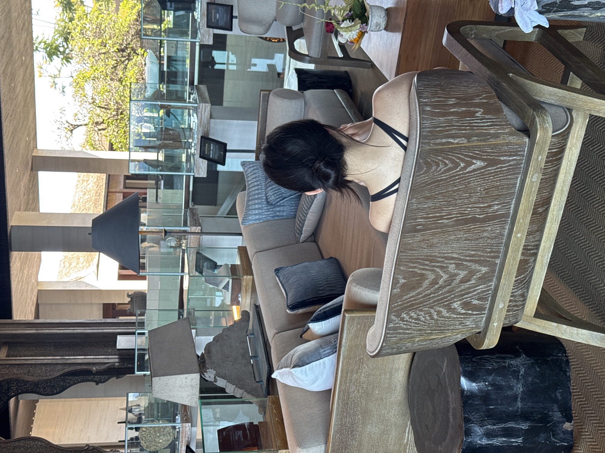 The image shows an interior space in Kuta Selatan, Indonesia. A woman with dark hair pulled into a bun is seated in a wooden chair. She's wearing a black top with spaghetti straps. The chair is part of a set with another identical chair. The space features a neutral-toned couch with various throw pillows and a dark marble side table. There's a glass display case that reaches high and allows visibility outside to trees and sunlight. Other furniture is visible in the background.