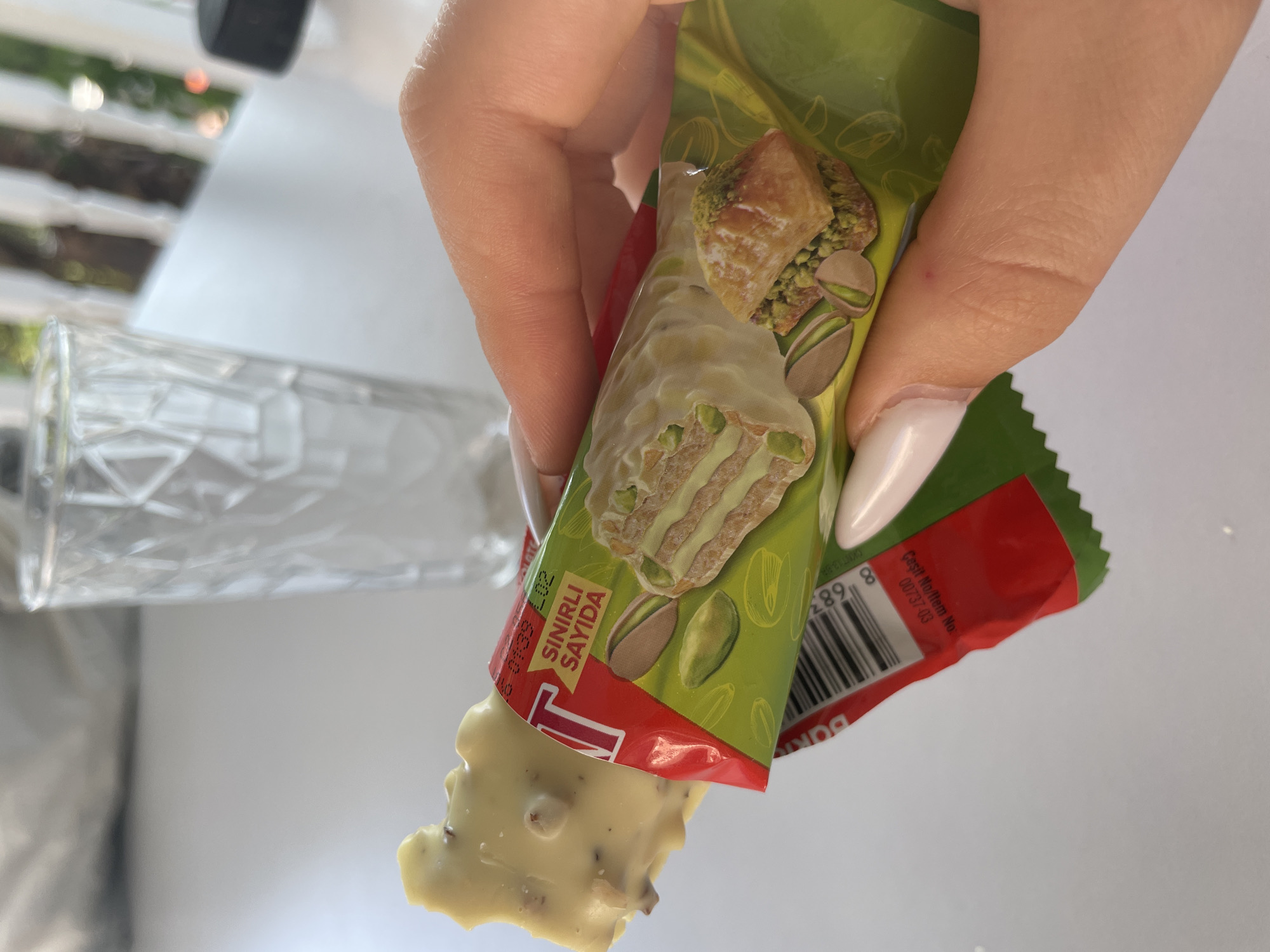 In Skala Avdiron, Greece, a hand with long, manicured nails holds a partially eaten white chocolate bar with pistachios. The bar is wrapped in green packaging that features an illustration of a piece of baklava and pistachios. The packaging also displays the text "SINIRLI SAYIDA" and a barcode. In the blurred background, a clear glass with a textured pattern is visible on a white surface.