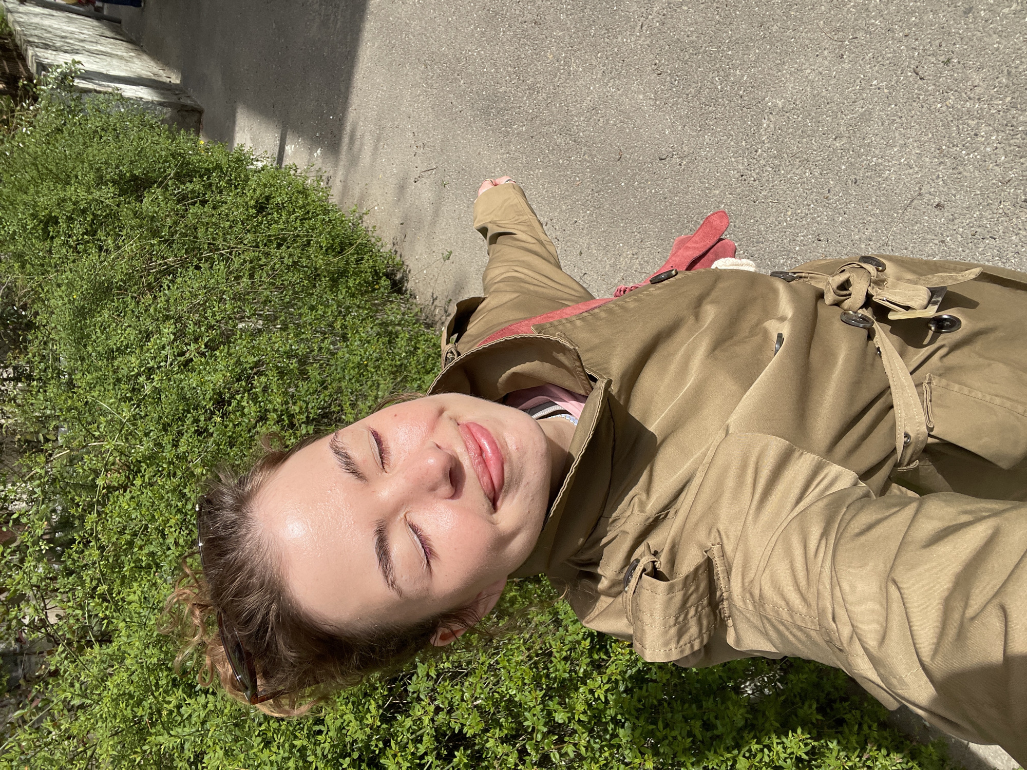 A young woman with closed eyes and a slight smile poses for a selfie outdoors, tilted to the left. She wears a tan trench coat over a pink top, and her hair is pulled back, revealing sunglasses tucked behind her ear. She stands beside a patch of green shrubs, with a gray sidewalk extending beyond her. The background features a sunlit building, suggesting the photo was taken during daylight. Based on the context provided, the location is likely Tbilisi, Georgia.