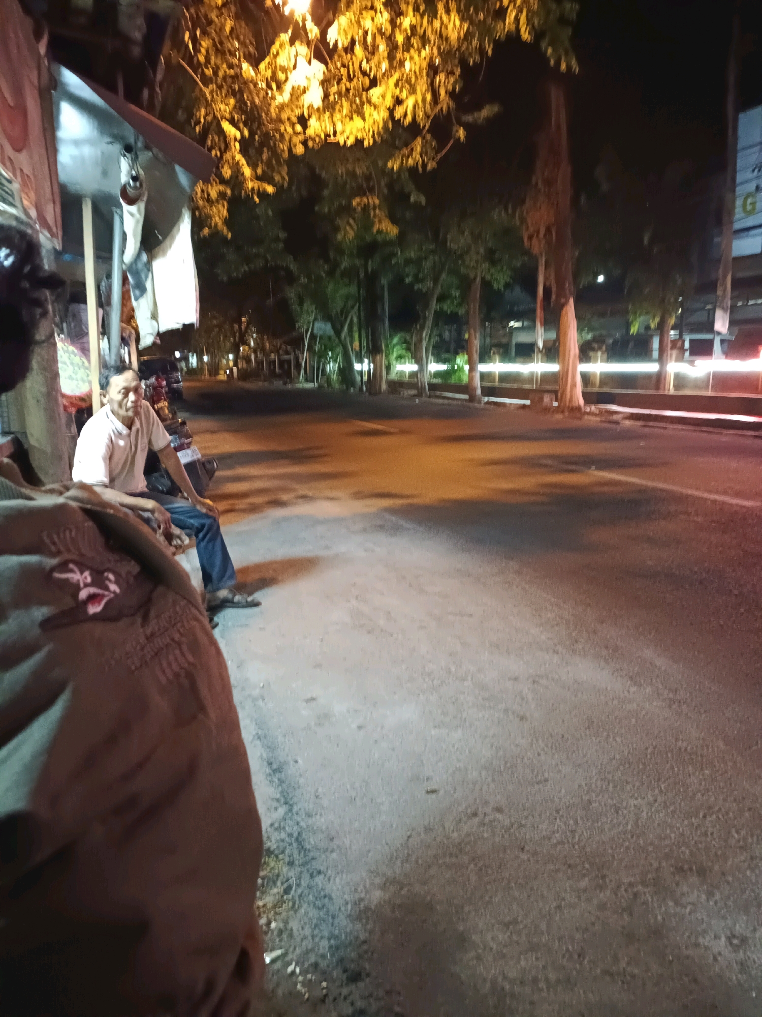 A medium shot captures a man sitting on a motorcycle parked beside a street stall at night. The man is wearing a light-colored polo shirt and blue jeans, with his hands resting in his lap. He is looking to his left. In the foreground, a person's shoulder and arm are visible, wearing a brown garment with an embroidered design. The background shows a street lined with trees and lights, suggesting an urban environment. The scene appears to be in Lamongan, Indonesia, given the context. The lighting is dim, with warm yellow and orange tones from streetlights and the stall's illumination.