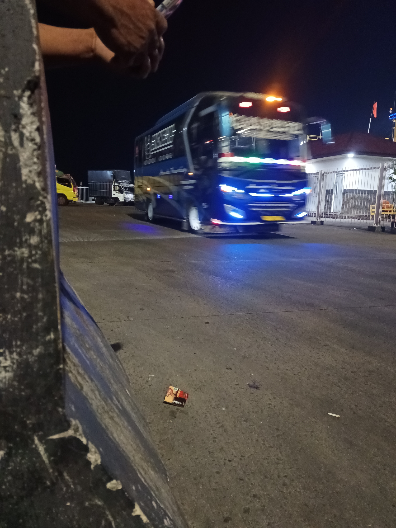 The image depicts a blurry nighttime scene at the Bakauheni ferry terminal in Indonesia. A blue bus with illuminated headlights and undercarriage lights is the primary subject, captured in motion. To the left, a yellow truck is partially visible. In the background, a white building with a fence and an orange flag on a pole is present. The ground is paved asphalt, with some debris, including a cigarette pack and a single cigarette butt, visible in the foreground. The overall atmosphere is dimly lit and dynamic due to the moving vehicles.
