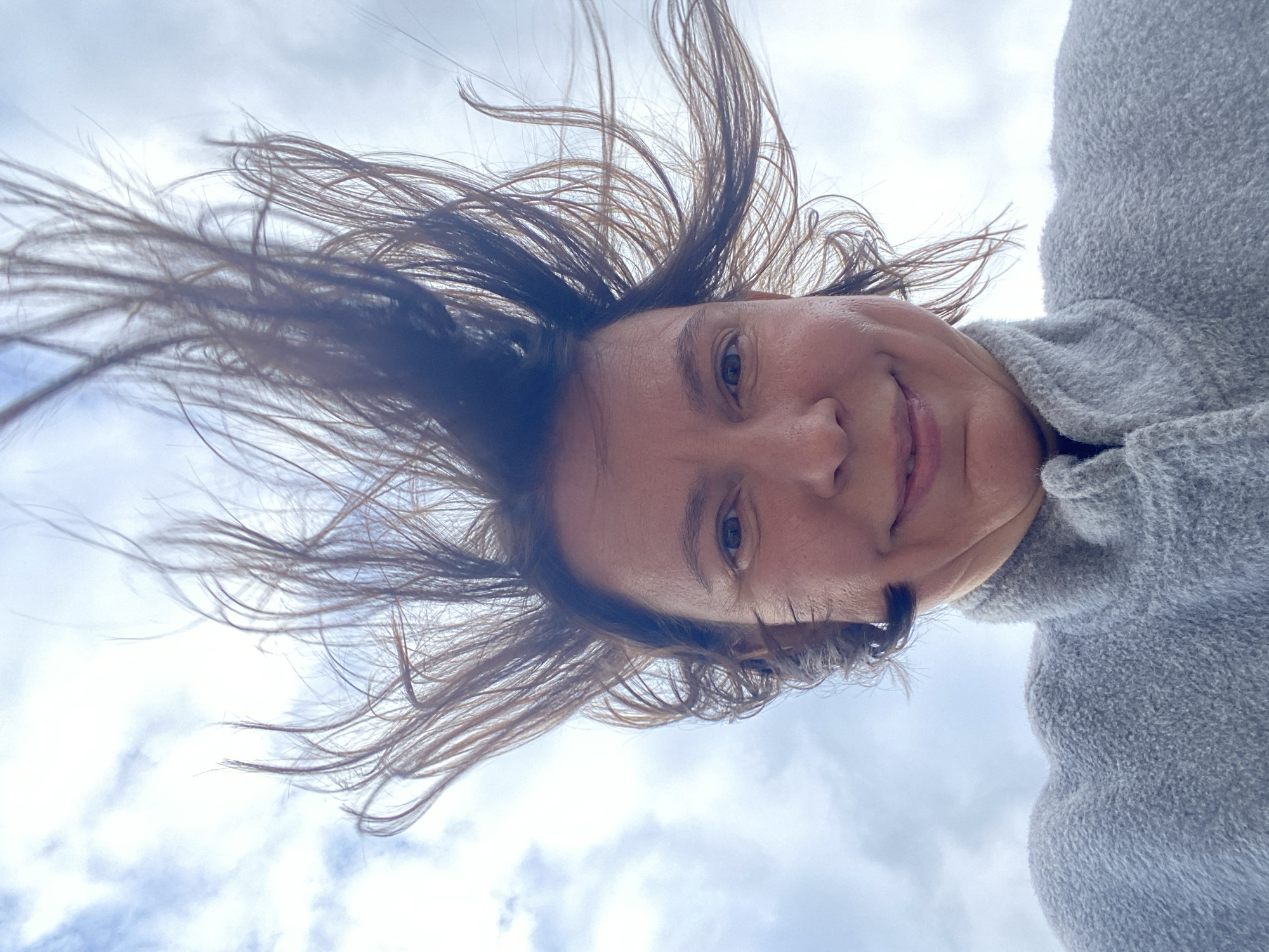 The image presents a close-up, low-angle portrait of a person against a cloudy sky in Chaikovsky, Russia. The main subject is a person's head, with their dark brown hair appearing dynamically splayed upwards and outwards, possibly due to wind or movement, creating an energetic impression. Their face is visible from the forehead to the mouth, with discernible dark eyes looking towards the viewer and a pleasant, gentle smile. The person is wearing a fuzzy, grey, high-collared garment, likely a warm sweater or jacket, which occupies the right side of the frame. The background is a pale blue and white cloudy sky, indicating an outdoor setting during daytime. No text is visible in the image.