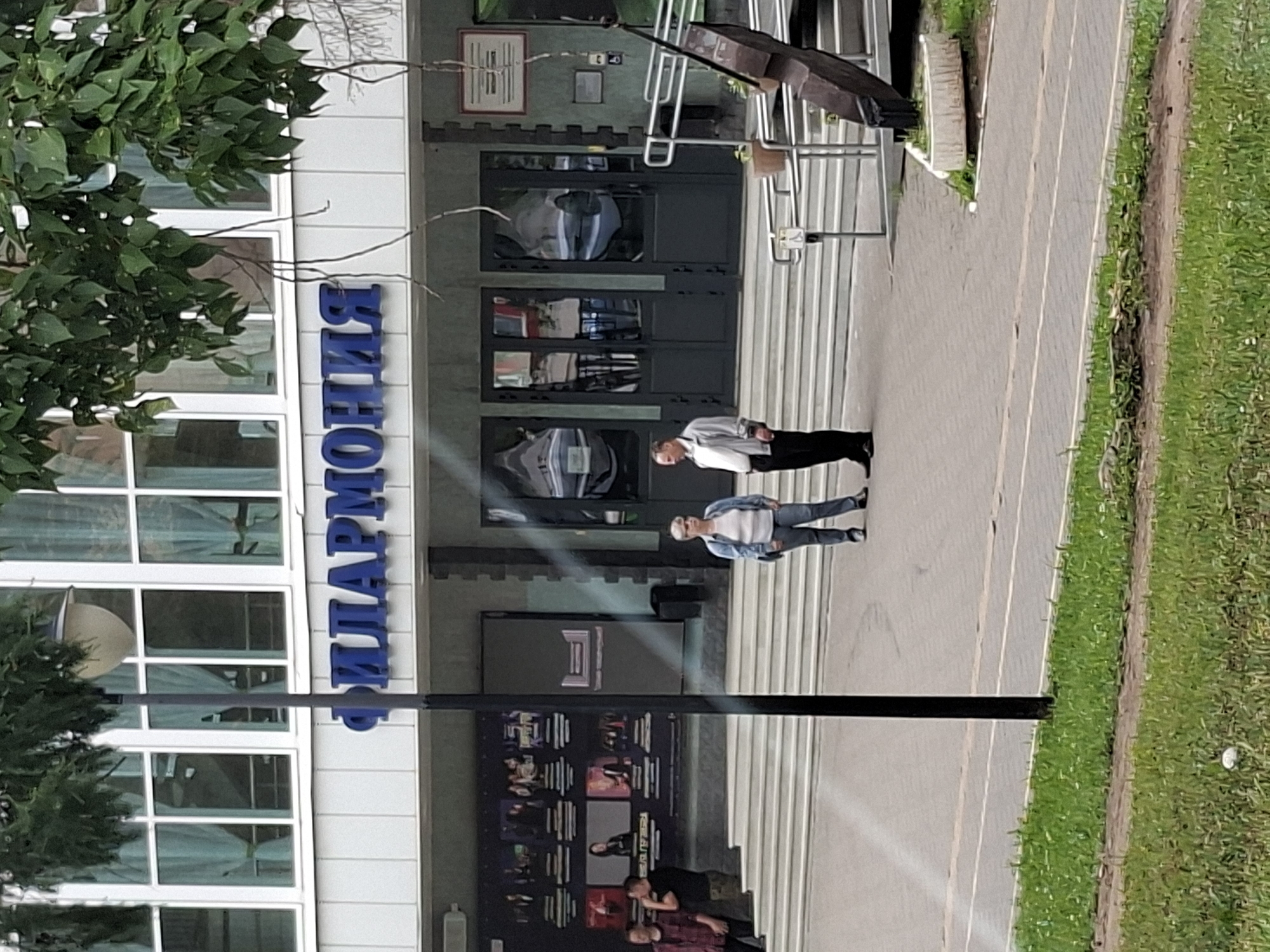 The image shows the Philharmonic Hall in Izhevsk, Russia, an architectural building with large windows and blue lettering spelling "ФИЛАРМОНИЯ" (Philharmonic) above the entrance. Two people, a man and a woman, are standing on the steps in front of the building, seemingly engaged in conversation. The man is wearing a light-colored jacket and dark pants, while the woman is dressed in a denim jacket and jeans. To the left of the entrance, a poster displays images of various performers. The foreground features a grassy area with a few scattered white objects, possibly flowers or litter, and a paved pathway. The scene appears to be a public space on an overcast day.