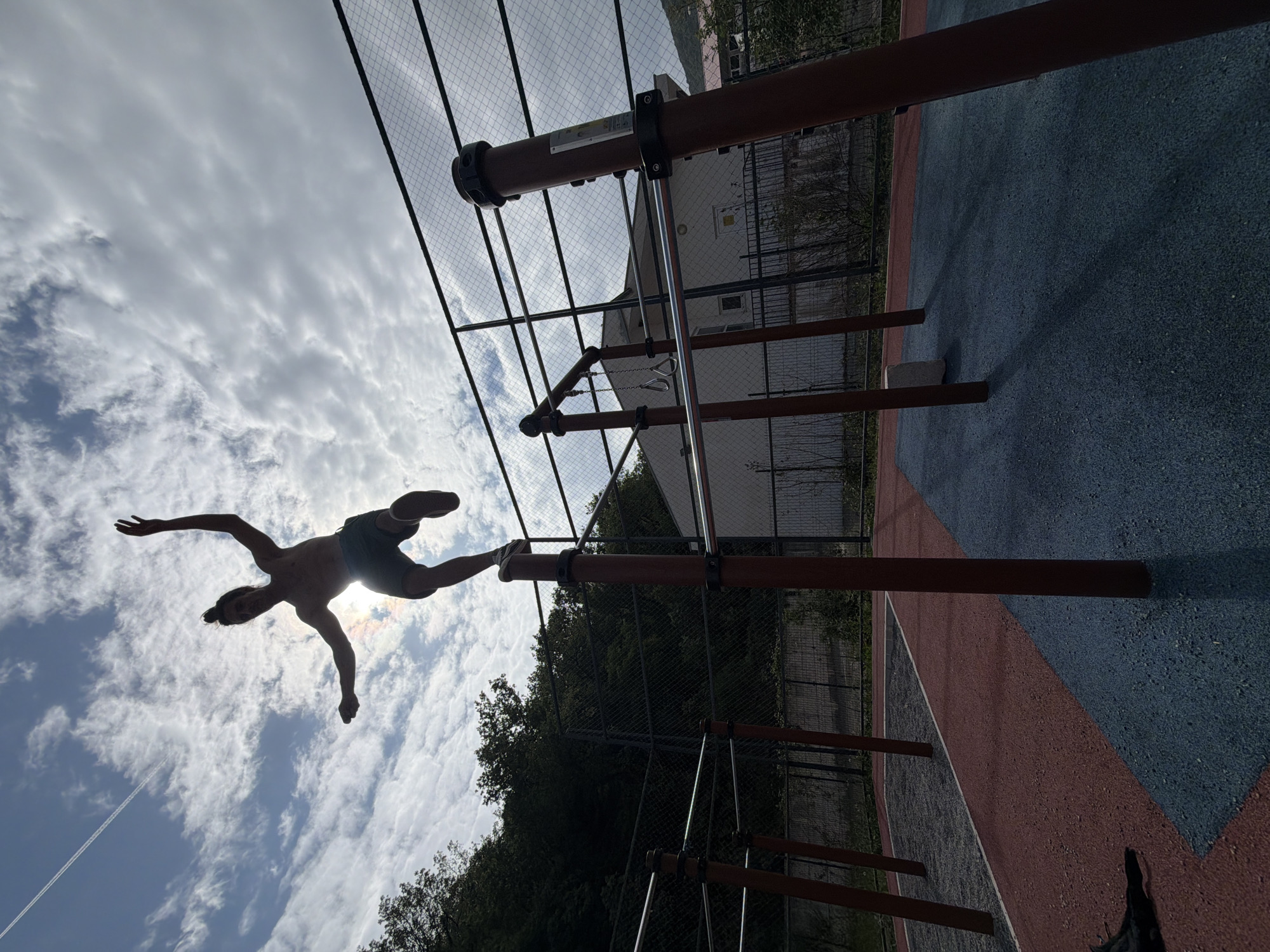 A person in shorts is suspended from a horizontal bar at an outdoor exercise park with both hands and one foot, silhouetted against a cloudy sky and the sun. The park, located in Petrovac na Moru, Montenegro, features brown metal bars and a safety net fence. The ground is colored red and blue. Trees and a building are visible in the background.
