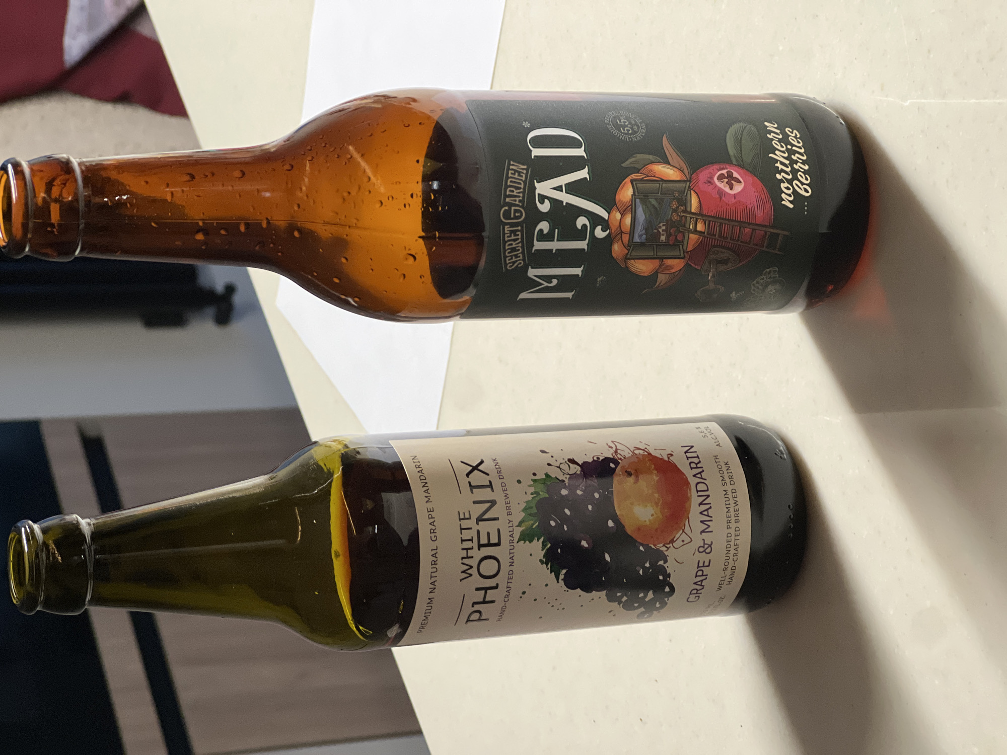 The image shows two bottles laying on a white surface. The top bottle is amber-colored and has a label reading "SECRET GARDEN MEAD" with the subtitle "northern berries." The bottle below is green and has a label reading "WHITE PHOENIX" with the subtitle "GRAPE & MANDARIN." There is a piece of white paper behind the top bottle. It is unclear if this photo was taken in Рубцово, Russia.