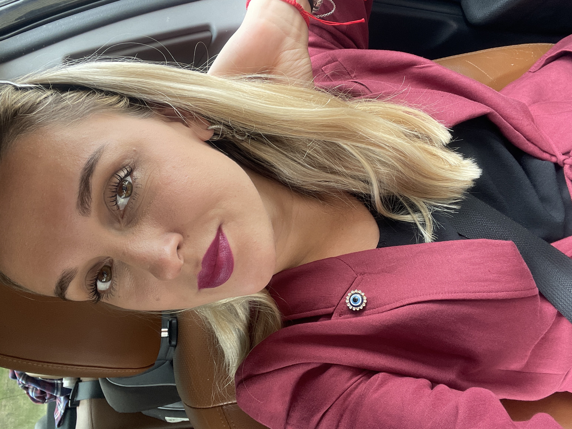 A blonde woman is sitting in the passenger seat of a car, her body angled towards the camera. She is wearing a maroon blazer over a black top, and a black seatbelt is visible across her chest. Her left arm is bent, and her hand is resting on her head, adorned with a red string bracelet and a silver bracelet. She has on a small, circular brooch with a blue and white evil eye design on the lapel of her blazer. Her makeup is prominent, with dramatic eye makeup and dark lipstick. Her hair is long and blonde, with some strands falling across her face. The interior of the car is visible, with brown leather seats and part of the dashboard. The lighting suggests it is daytime.