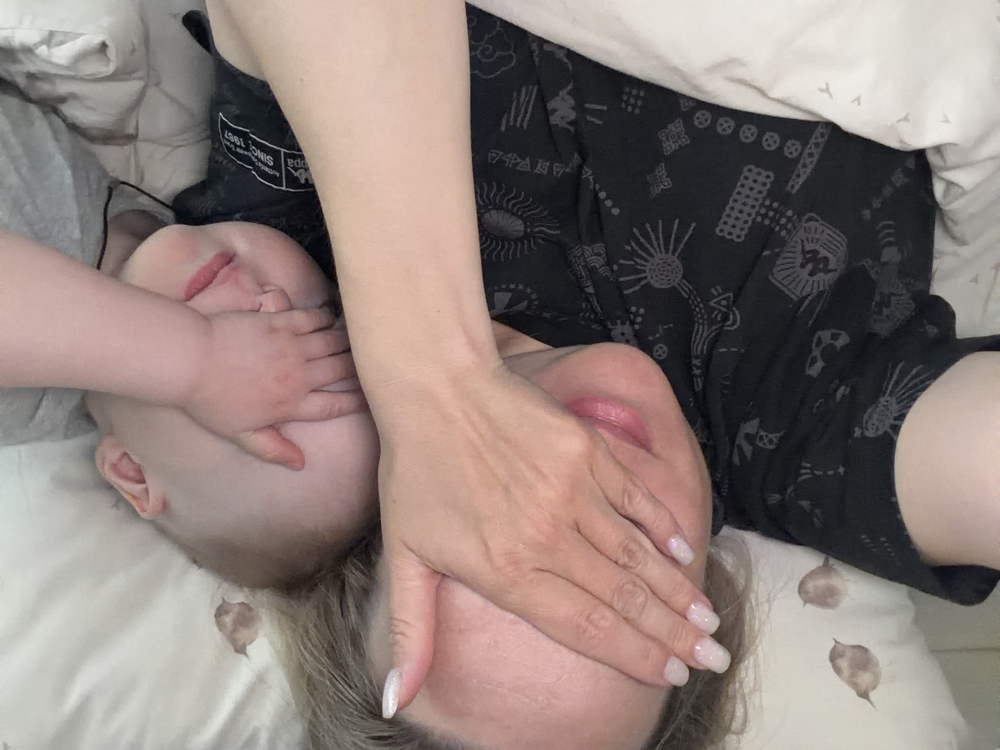 An overhead shot captures an adult and a child lying closely together on a bed, engaged in a playful or affectionate interaction. The adult, positioned on the right side of the frame with their head towards the bottom, has light-colored hair and wears a dark-patterned garment. Their right hand, adorned with clear, shimmering nail polish, covers both their own eyes and partially the child's face. A gentle smile is visible on the adult's lips.
The child, likely a baby or toddler, is on the left side, also with their head towards the bottom. Their small hand is similarly placed over their own eyes and mouth, overlapping with the adult's hand. Both individuals have light skin tones. The child appears to be wearing light-colored clothing, possibly gray.
The bedding is white or light-colored, with subtle brown, leaf-like patterns visible on the pillow near the adult's head. The adult's dark garment features various abstract and sun-like designs. A label on the adult's clothing is visible, showing the text "KAPPA" and "SINCE 1967". The overall scene suggests a moment of warmth, playfulness, or rest, likely indoors in a bedroom in Votkinsk, Russia.