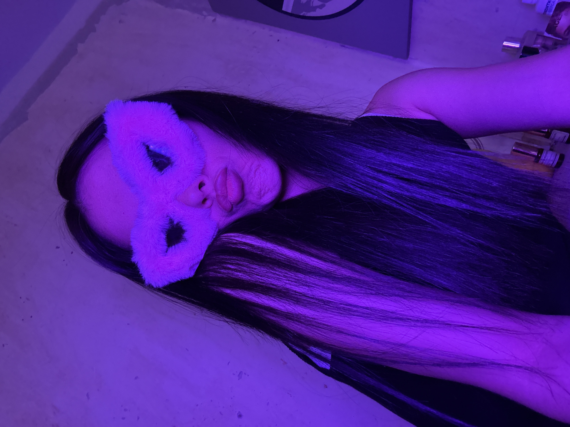 A young woman with long, dark hair, featuring some pink-purple streaks, is captured in a close-up selfie. She is wearing a fluffy pink eye mask that resembles cat ears and is puckering her lips. The lighting in the image is predominantly purple, suggesting an artificial light source, possibly a colored bulb or LED strip. The background appears to be a textured wall. On the right side of the image, there are some objects on a shelf, including what appear to be bottles, possibly of perfume or other cosmetics, and a framed picture of a person. The overall scene suggests a personal, indoor environment, potentially a bedroom or vanity area. The location is specified as Votkinsk, Russia.