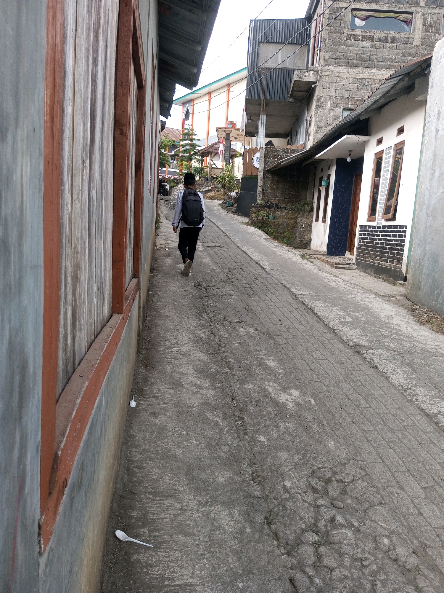 A narrow alleyway in Kalibeber, Indonesia, is shown in this full-frame, eye-level shot. On the left, a weathered wooden wall with red trim lines the path. In the distance, a person wearing a white long-sleeved shirt, black pants, and a black backpack walks away from the camera. The alleyway is paved with grey bricks, and the surrounding buildings vary in architectural style, with one featuring a white facade and black brick accents, and another constructed from grey cinder blocks. The scene is overcast. Two white plastic spoons are visible on the ground.