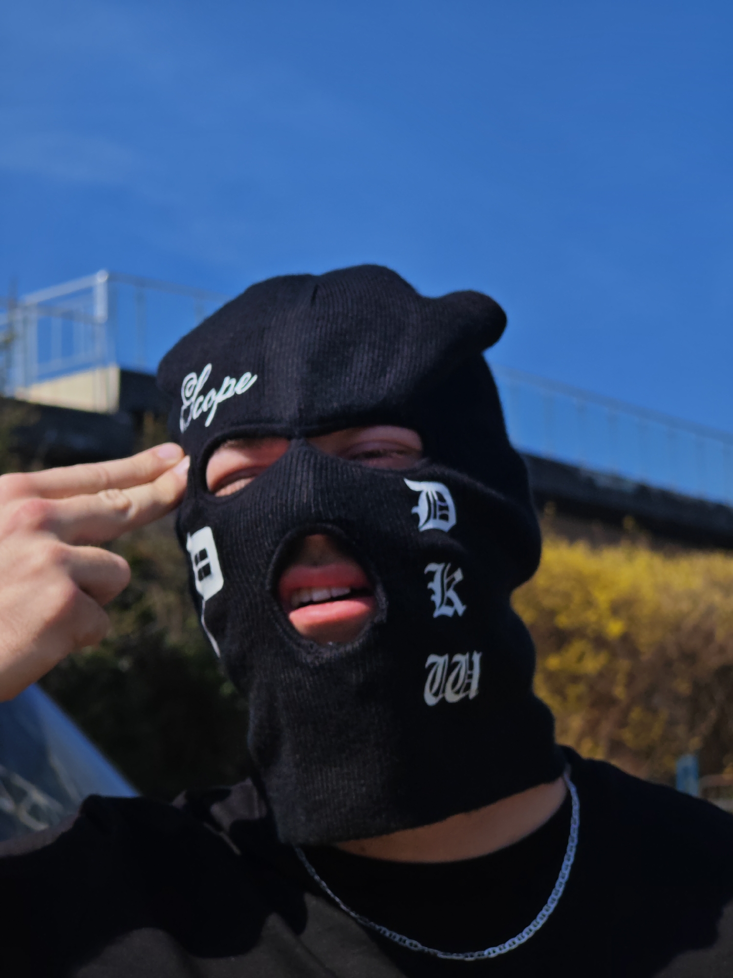 The image depicts one individual outdoors in Shumen, Bulgaria, under a clear blue sky, suggesting daytime. The subject is partially visible from the chest up, wearing a black knit balaclava with white embroidered text: "Scope" across the forehead and stylized letters resembling "D", "K", and "WH" vertically aligned on the wearer's right cheek. The eyes and mouth are visible through the balaclava's openings; the mouth is slightly open, revealing teeth and tongue. The individual is also wearing a black t-shirt and a thin silver-colored chain necklace. The subject's right hand is raised to their temple, performing a "finger gun" gesture with the index finger and thumb extended. In the blurred background, a light-colored structure with blue accents is visible to the upper left, while yellow foliage is present to the upper right. A dark, reflective surface, possibly part of a vehicle, is partially visible in the lower-left foreground.