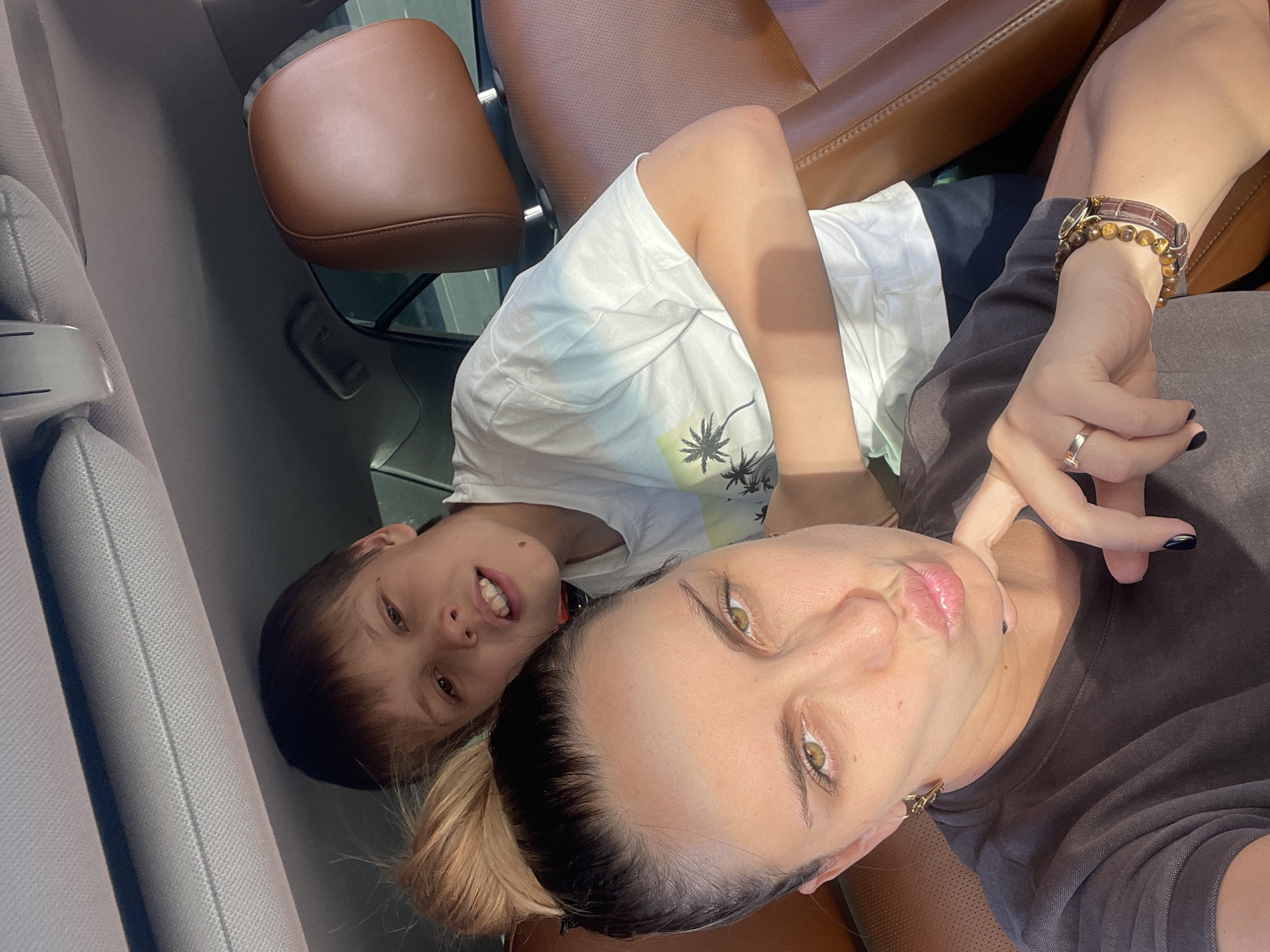 Two people, a woman and a boy, are visible in the image. They appear to be inside a car, given the leather seats and interior car features. The woman is holding the camera, taking a selfie with the boy. The boy is lying down, looking at the camera with a slight smile. The woman is also looking at the camera. No specific city information is discernible from the image.