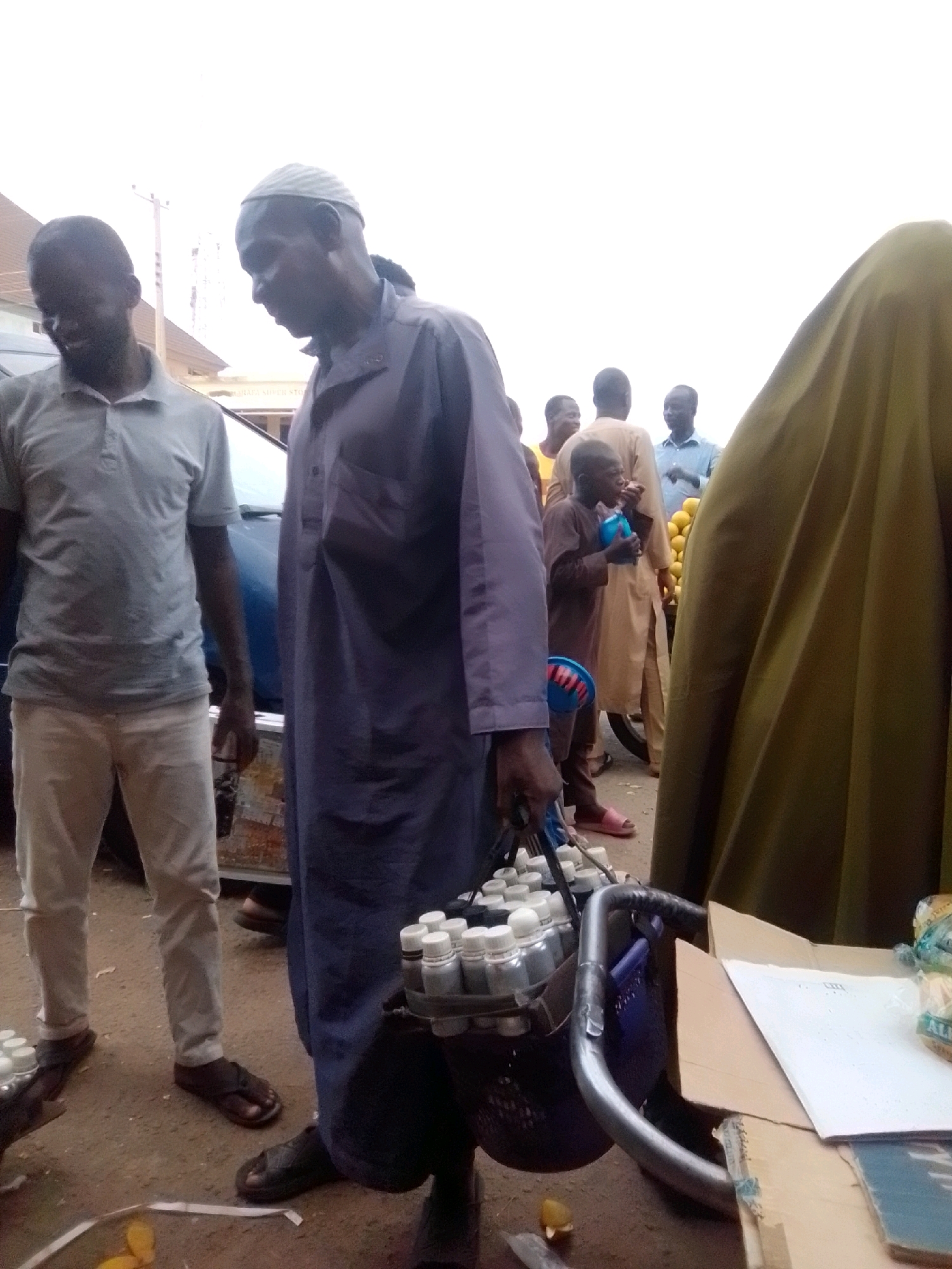 In the city of Arawa, Nigeria, a group of people are gathered outdoors, possibly at a market. A man in a purple robe and a white cap is the central figure, carrying a black basket filled with small bottles on his right arm. He is looking down at the basket. To his left, a man in a grey polo shirt and light-colored pants stands, looking to his left. Behind the central figure, several other individuals are visible, including a young boy in a brown outfit holding a blue container and eating. A woman in a large, olive-green head covering is positioned to the far right of the frame. The ground is dirt, and there are some orange fruits scattered around. A blue car is partially visible behind the two main figures. The sky is overcast.