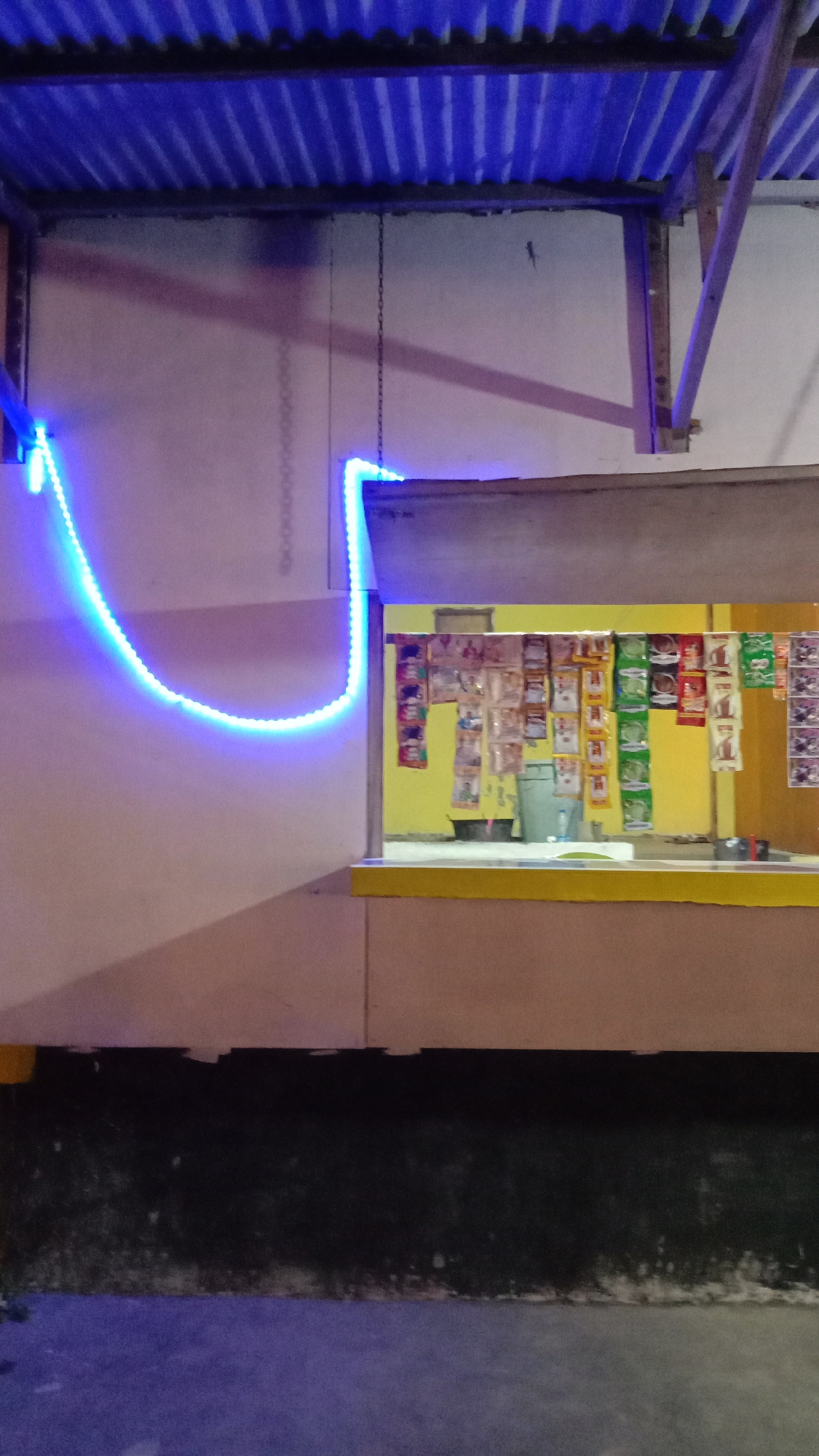 The image captures an illuminated small shop or stall at night. A prominent blue LED strip light traces a wavy path across a light-colored exterior wall on the left and also outlines the top and side edge of a service window or counter. Above, a corrugated metal roof is visible, with its underside reflecting a blue hue from the artificial lights.
Through the shop's rectangular opening, a bright yellow interior wall is seen, decorated with rows of various small, individually packaged items such as sachets or pouches, likely containing snacks, drinks, or groceries. A dark bucket or bin is also visible inside the shop. To the right, a wooden or metal support beam for the roof structure is present. A thin metal chain hangs from the roof support. A small lizard can be discerned on the light-colored wall above the blue LED strip, near the support beam. The foreground shows a dark ground surface, possibly pavement or dirt.
The scene suggests a modest convenience store or warung, characteristic of establishments found in places like Lhokseumawe, Indonesia, open or recently closed for the evening. There are no people visible, and no discernible text on any objects.