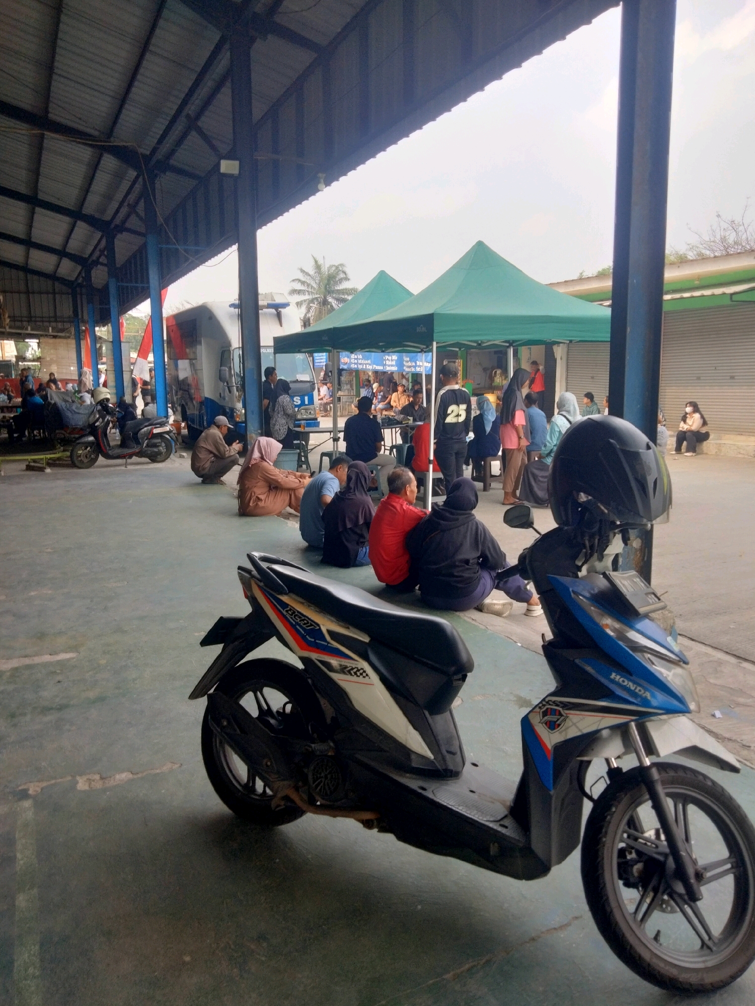 The image depicts an outdoor scene in Sukaasih, Indonesia, under a covered structure with a corrugated metal roof supported by blue and dark-colored pillars. In the foreground, two scooters are parked, one prominently featuring a black helmet on its handlebars. The ground is paved with concrete. In the middle ground, several people are seated on the ground, appearing to be waiting. Some are facing away from the camera, while others are in profile. Two green tents are set up in the background, one behind the other. A white truck with "POLISI" (Police) written on its side is visible behind the tents. Further back, more people are gathered, and some buildings with metal roll-up doors can be seen. The sky is overcast. The overall atmosphere suggests a public gathering or waiting area.