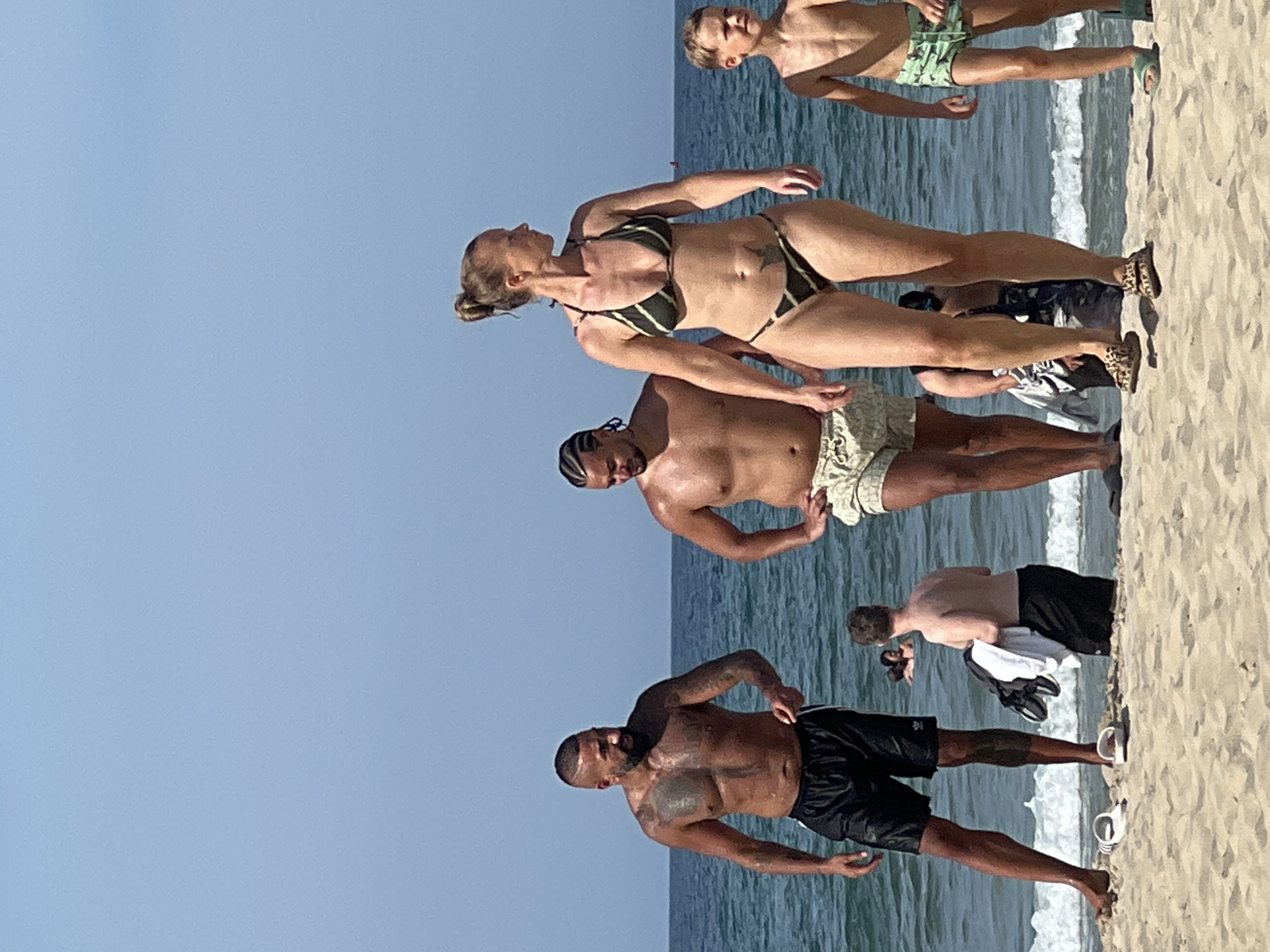 The image shows a group of people on a sandy beach. The sun is shining brightly, suggesting it is daytime. The water of the ocean is visible with waves breaking on the shore.
In the foreground, a man with tattoos on his torso and arms is wearing black swim shorts. To his left, another man is wearing light-colored patterned swim trunks.
Further back, a woman in a striped bikini is standing. Behind her, a young boy is looking towards the water.
The scene is set on a beach, a popular location for recreation and relaxation, especially on a sunny day. The presence of the ocean and sand indicates an outdoor environment. The people are dressed in swimwear, suggesting they are enjoying a beach day, possibly swimming or sunbathing. The overall atmosphere appears casual and relaxed. No visible text is present in the image.