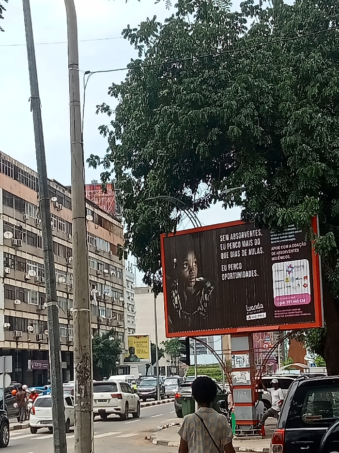 This daytime image captures a lively urban street scene in Luanda, Angola, characterized by multi-story buildings, dense foliage, and active traffic. In the foreground, a person with dark hair, wearing a light-colored top, is seen walking away from the camera on a sidewalk, alongside utility poles and overhead wires.
A large, eye-catching billboard dominates the mid-right of the frame, partially framed by a large tree with abundant green leaves. The billboard displays a social awareness message in Portuguese, featuring a solemn black-and-white image of a child. The text on the billboard reads: "SEM ABSORVENTES, EU PERCO MAIS DO QUE DIAS DE AULAS. EU PERCO OPORTUNIDADES." (Without sanitary pads, I lose more than school days. I lose opportunities.) Below this, it urges: "APOIE COM A DOAÇÃO DE ABSORVENTES HIGIENICOS." (Support with the donation of hygienic sanitary pads.) The billboard also includes the word "Luanda," "Presidente," and various corporate/organizational logos, along with a contact number: "957 443 434."
The street itself is busy with several cars, including a white SUV, suggesting an active urban environment. Multi-story buildings line the street, many featuring numerous air conditioning units and satellite dishes on their facades. Further down the street on the left, a smaller billboard is partially visible with text that includes "A RAINHA..." (The Queen...). The sky appears overcast, indicating a cloudy day. Several indistinct figures of people are visible on the sidewalks and near the vehicles, contributing to the bustling street atmosphere. A green trash bin is also partially visible on the sidewalk.