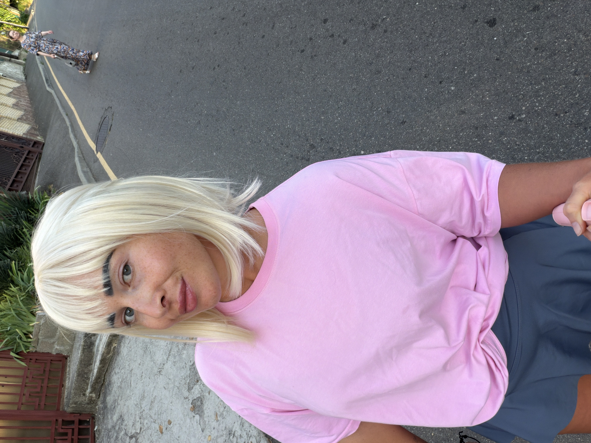 In the city of Sochi, Russia, a woman with blonde hair and distinctive dark eyebrows poses for a selfie on a paved street. She is wearing a pink t-shirt tied at the waist and a grey skirt. The background shows a sidewalk, a building with a red gate, and lush greenery. Further down the street, another woman in a patterned dress walks away, pulling a small suitcase. The scene appears to be outdoors during daylight.