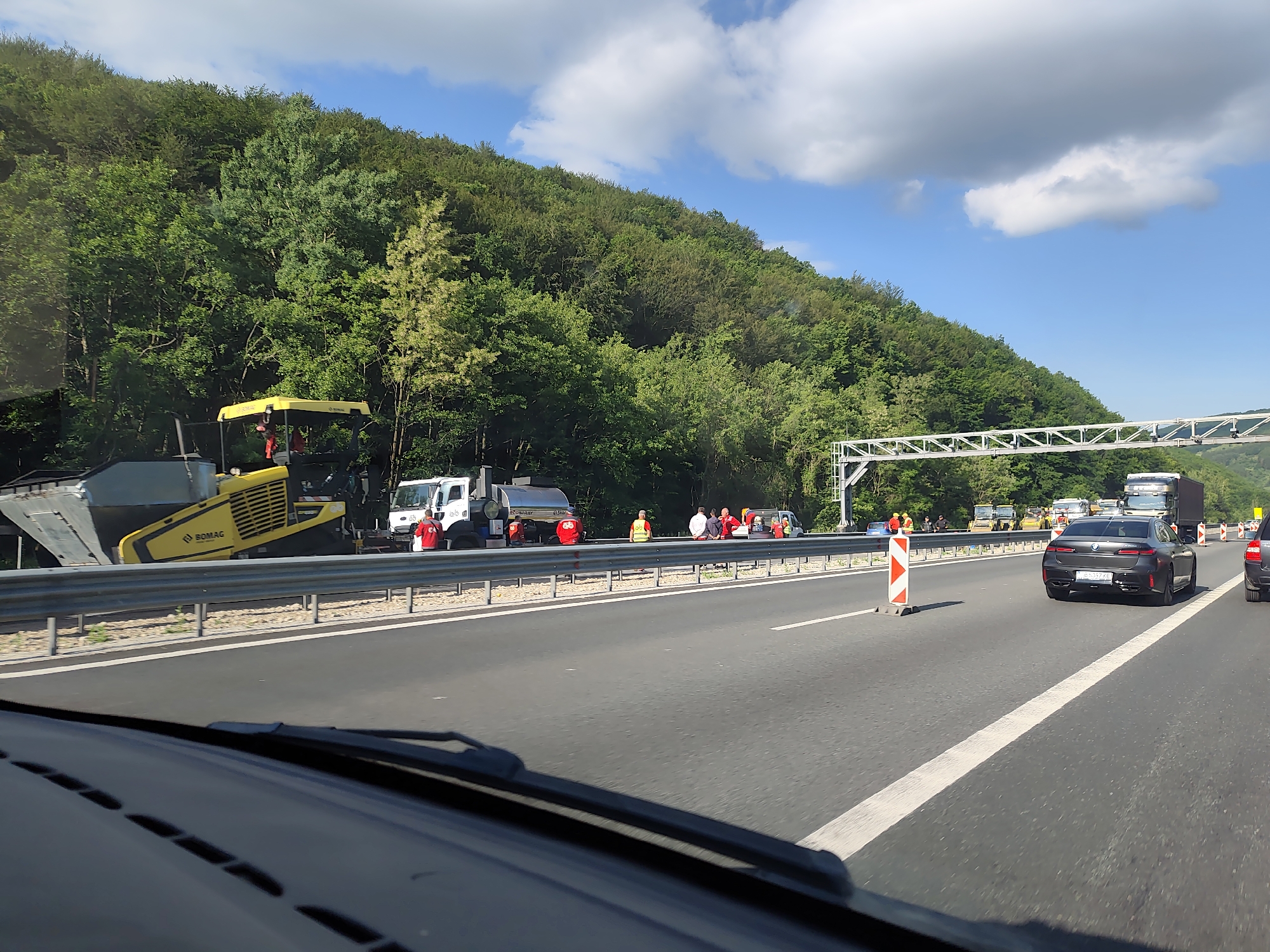 The image depicts a construction scene on a highway near Churek, Bulgaria. In the foreground, the dashboard and windshield of a vehicle are visible, implying the photo was taken from a moving car. The highway itself is paved, with white lane markings.
On the left side of the highway, a yellow BOMAG asphalt paving machine is stationary. Behind it, a white truck with a silver tank is parked, and a white pickup truck is also present. Several individuals wearing bright red or yellow vests are gathered near these vehicles, likely engaged in road construction or maintenance. A large, wooded hillside forms the backdrop, densely covered in green trees.
Further down the highway, construction barriers and traffic cones are visible, indicating a work zone. Several cars and trucks are driving on the highway, with a dark-colored BMW sedan prominently featured in the middle lane. Traffic appears to be moving. The sky is blue with scattered white clouds. A large metal structure, possibly a gantry for traffic signs or sensors, spans the highway in the distance. The overall scene suggests ongoing road work on a sunny day.