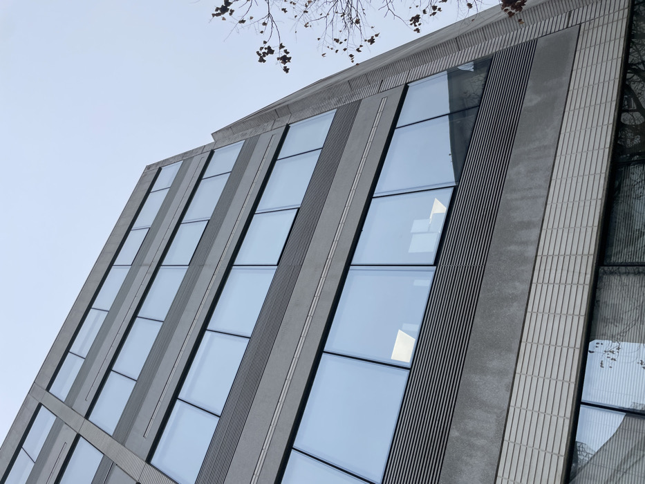An upward-angled view captures the exterior of a multi-story building located in Montigny-le-Bretonneux, France. The building presents a modern, rectilinear design featuring multiple levels. The facade is predominantly grey, composed of textured panels. Each floor displays a series of large, rectangular windows with dark frames, reflecting an overcast, light grey sky. Between these window bands, horizontal sections of grey louvers or slats are present. Below the louvers, a lighter-toned, horizontally patterned material forms a band at the base of the visible structure. Reflections in the windows reveal interior artificial lighting on some floors and, in the lowest visible windows, the sparse, bare branches of a tree, some retaining a few dry leaves. No individuals or discernible activities are visible.