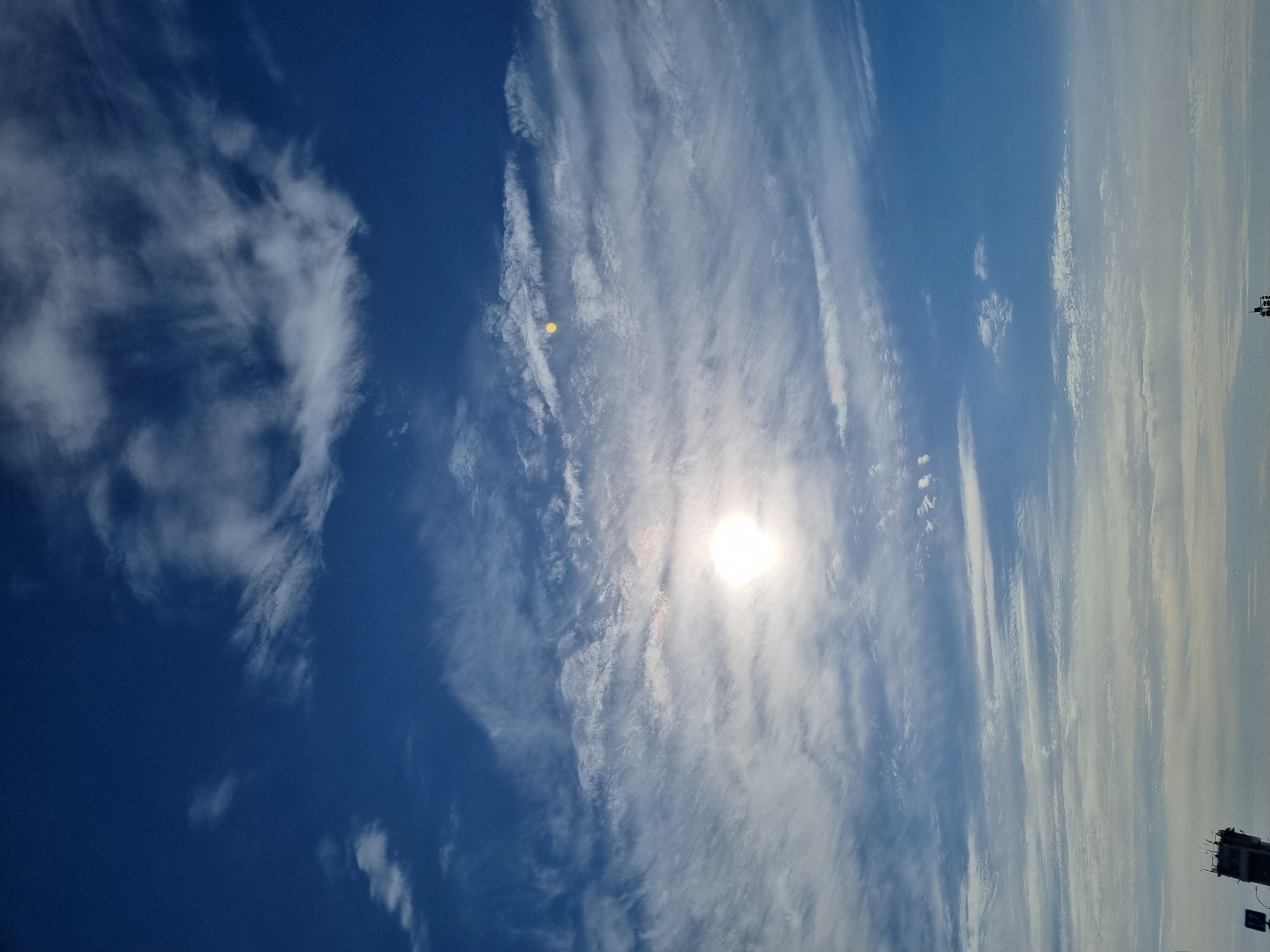 The image displays a daytime sky over Chaikovsky, Russia, dominated by a deep blue expanse interspersed with white, wispy cirrus and cirrocumulus clouds. The sun, a brilliant, overexposed white orb, is prominently positioned in the middle-left quadrant, creating a strong lens flare, with a distinct smaller yellow lens flare visible above and to its right. A faint, hazy horizon line is discernible at the very bottom of the frame. In the lower-left corner, a dark, multi-tiered structure with antenna-like elements on its roof, resembling a control tower or communication building, is partially visible, adjacent to a fragment of a blue and white sign. A smaller, indistinct dark structure is also present at the extreme bottom-right. No individuals, specific actions, or interactions are observable.