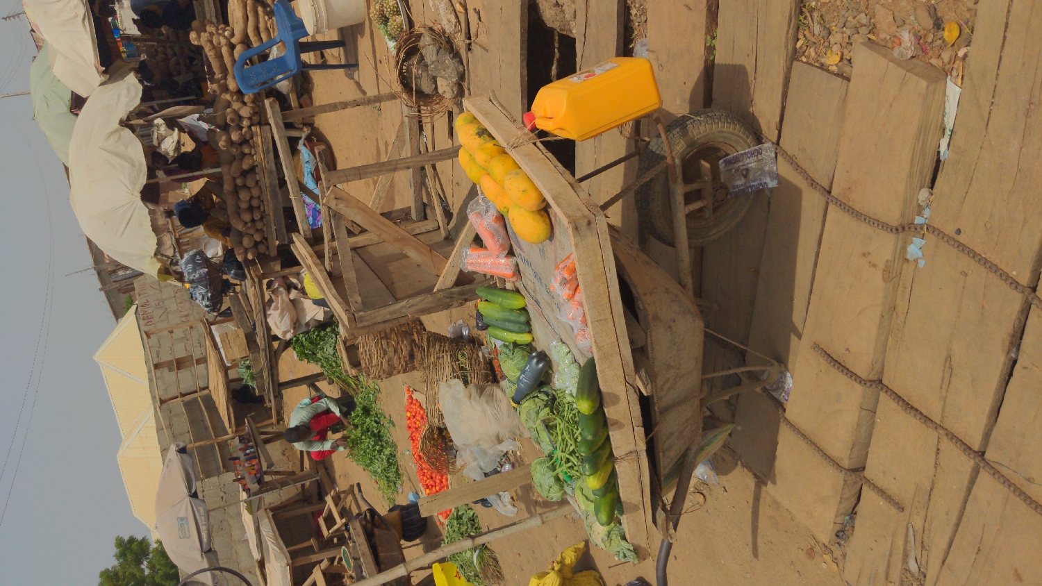 An outdoor market scene, likely in Ado, Nigeria, is captured from a slightly elevated perspective. The ground is unpaved dirt, with some wooden planks laid out.
In the foreground, a wooden cart serves as a display for a variety of fresh produce. On its top, ripe yellow papayas are stacked, and below them, plastic-wrapped carrots, green cucumbers, lettuce, and long green beans are arranged. A yellow jerrycan, possibly containing oil or water, rests on a wooden frame next to the cart, secured by a red string. A metal wheelbarrow with a tire, partially obscuring the text "OCHOLONA," is visible to the right. A blue plastic container nearby also shows "OCHOLONA."
To the left and behind the cart, several people are engaged in market activities. One individual, dressed in a red and blue top, crouches on the ground, sorting leafy green vegetables. Other vendors are partially visible under large white umbrellas, which provide shade from the bright daytime sky. Further back, another stall displays large piles of yams or cassava tubers, with a blue plastic chair and a white bucket nearby. Various woven baskets, plastic bottles, and bags are scattered around. The background shows modest buildings, some constructed with concrete blocks, under a pale, clear sky. The overall scene depicts a bustling, informal marketplace during the daytime.