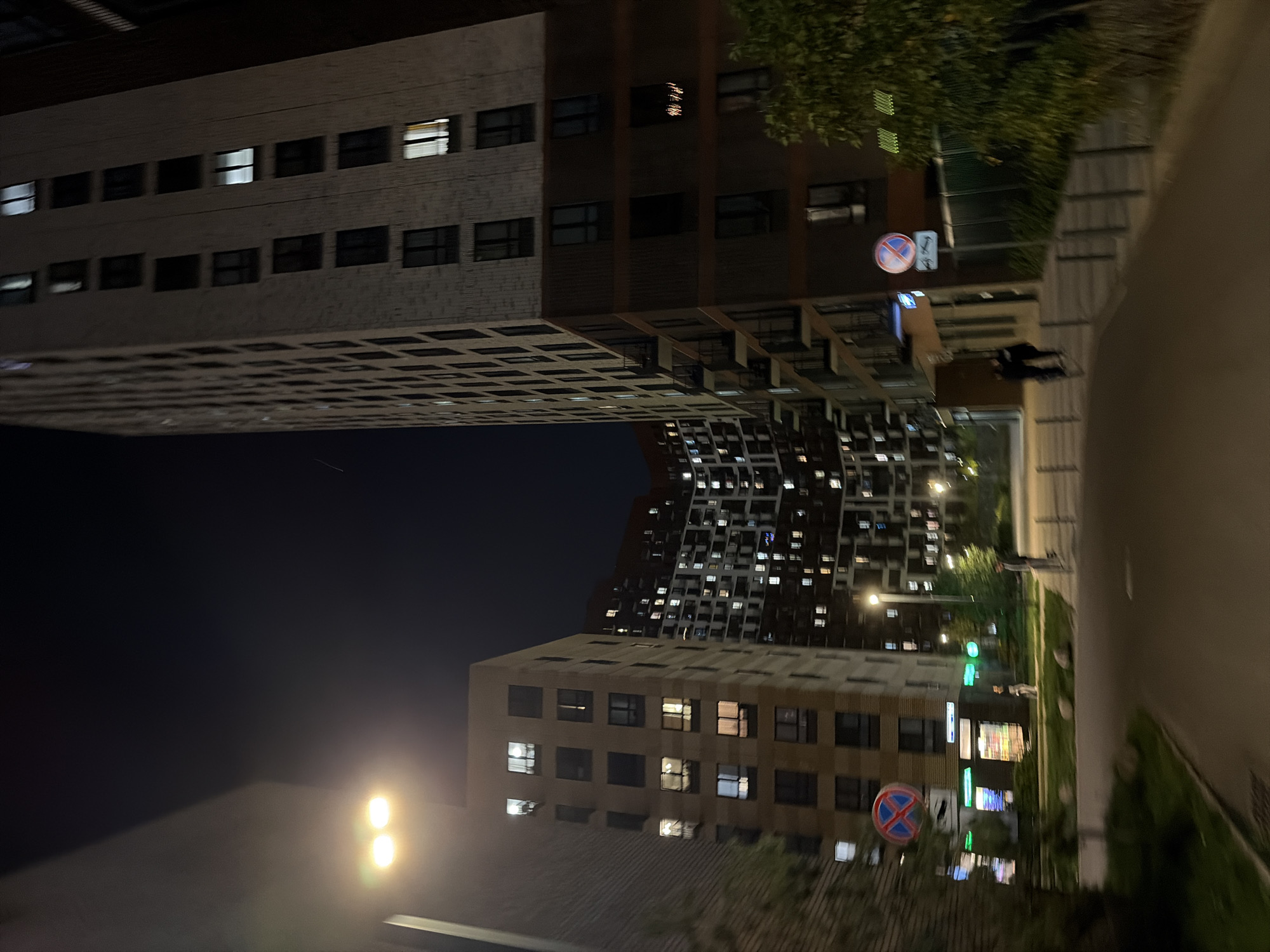 This image depicts a nighttime view of a modern residential complex in Moscow, Russia. The scene is dominated by tall, multi-story buildings with numerous illuminated windows, indicating occupied apartments. The architectural style suggests contemporary urban development. In the foreground, a paved pathway curves through the complex, bordered by low green landscaping and bollards. Streetlights illuminate the pathway and the facades of the buildings. A traffic sign, likely a "no parking" sign, is visible on the left side of the pathway. Several individuals are present, walking along the pathway. The overall impression is one of a well-lit, active urban environment at night.