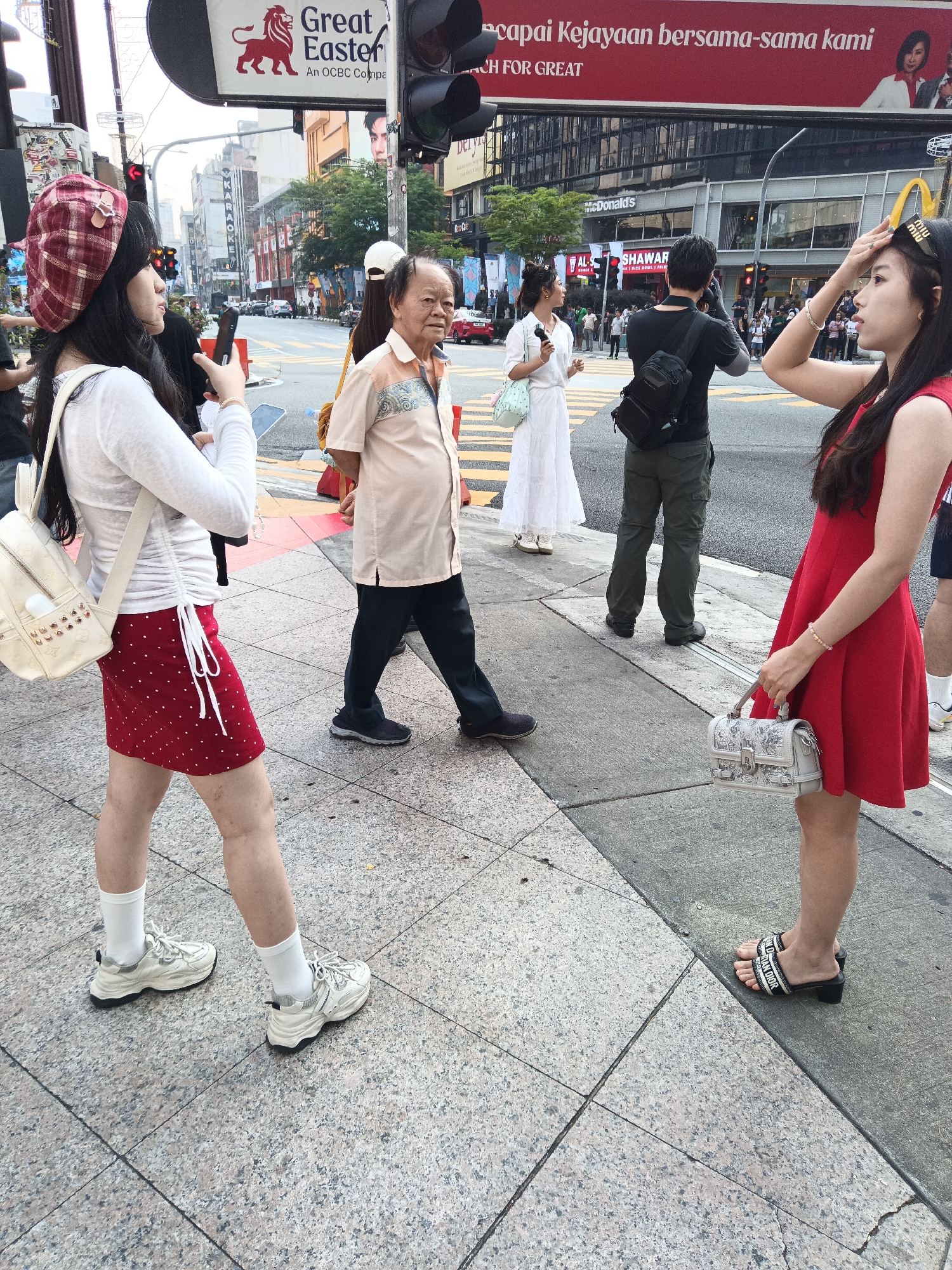 In Kuala Lumpur, Malaysia, on a busy street, a young woman wearing a red plaid beret, a white long-sleeved shirt, and a red polka-dot skirt is holding a smartphone. She has a light-colored backpack slung over her shoulder. To her right, an older man in a light-colored patterned shirt and dark pants stands facing forward. Further to the right, a woman in a white dress walks away from the viewer, holding a phone and a light-colored handbag. Another woman in a red dress stands with her back to the camera, holding a white handbag. The scene is set on a city sidewalk with a crosswalk visible, and a large billboard for "Great Easter" and "OCBC Bank" is overhead. Traffic lights and buildings line the street, indicating an urban environment.