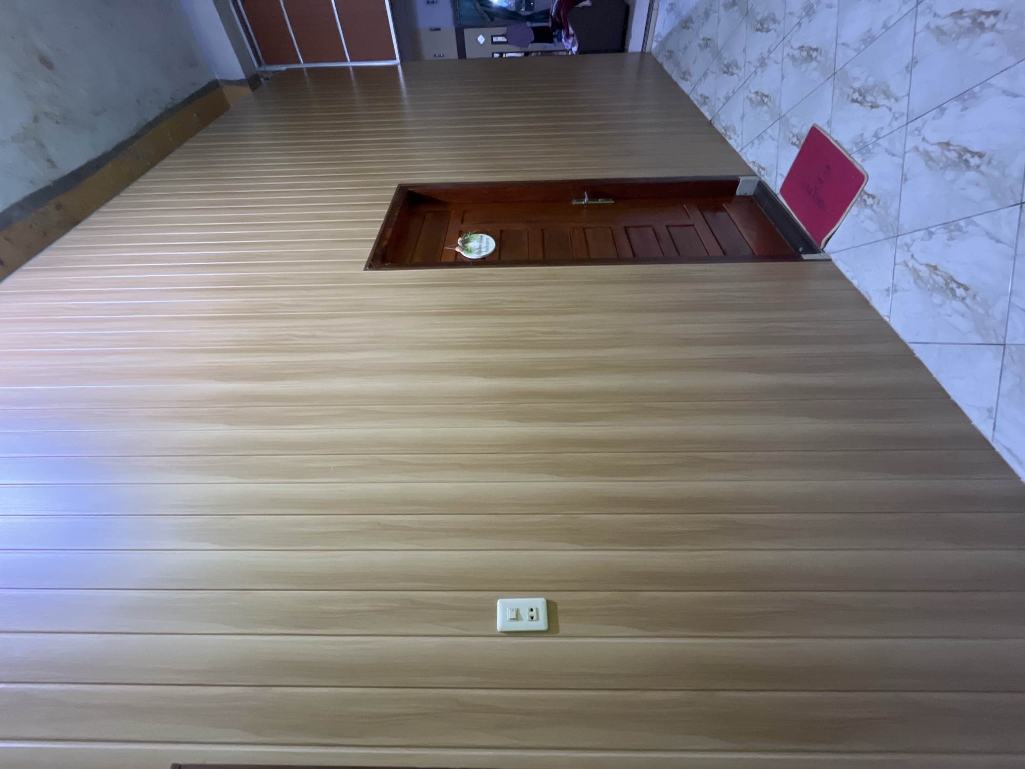 The image shows an interior wall, featuring a brown wooden door, a red doormat, and a light switch. The wall is clad in light brown, horizontally-aligned planks. The scene appears to be indoors in Lhokseumawe, Indonesia. A section of tiled floor is visible in the upper right corner.