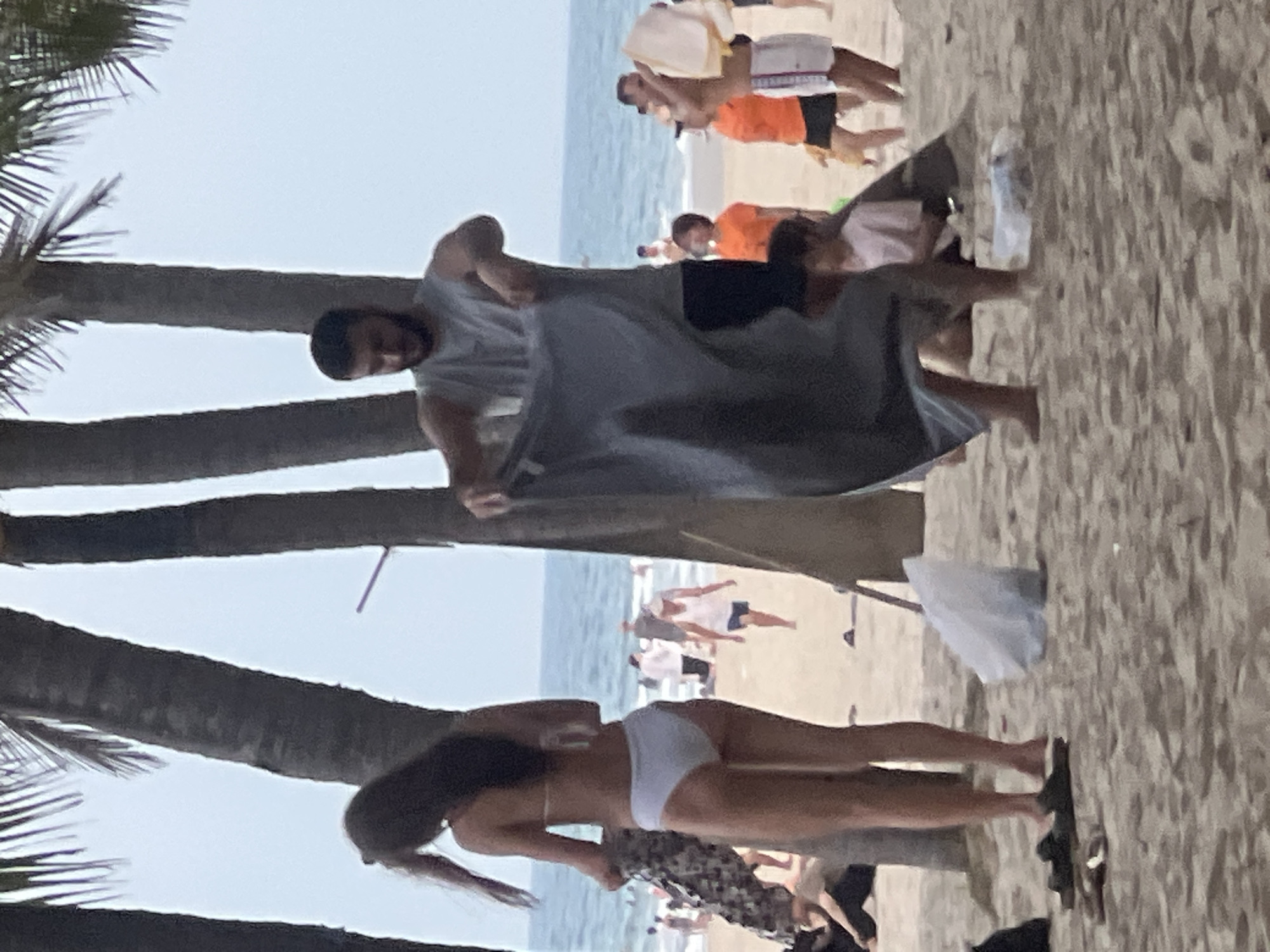 The image depicts a beach scene in Đà Nẵng, Vietnam, with several people visible. In the foreground, a woman in a white bikini is standing with her back to the camera, holding a patterned garment. To her right, a man wearing a grey tank top and black shorts is unfolding a large grey towel. Behind them, various individuals are scattered across the sandy beach, some walking and others sitting. The ocean is visible in the background under a hazy sky, with palm trees framing the scene. There are also a few plastic bags on the sand.