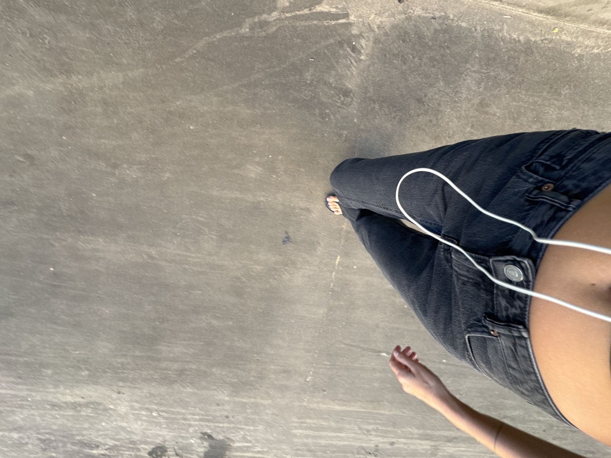 The image shows a person's legs and arm from a high-angle perspective, standing on a concrete surface. The person is wearing dark wash denim jeans and appears to be wearing flip-flops. White headphone cords are draped around the jeans. A person's arm is visible on the left side of the frame, with the hand extended. The concrete surface is textured and appears to be outdoors, possibly a walkway or street. There are some dark smudges and lighter streaks on the concrete. The image does not contain any discernible landmarks or details that would indicate the location is Moscow, Russia.
