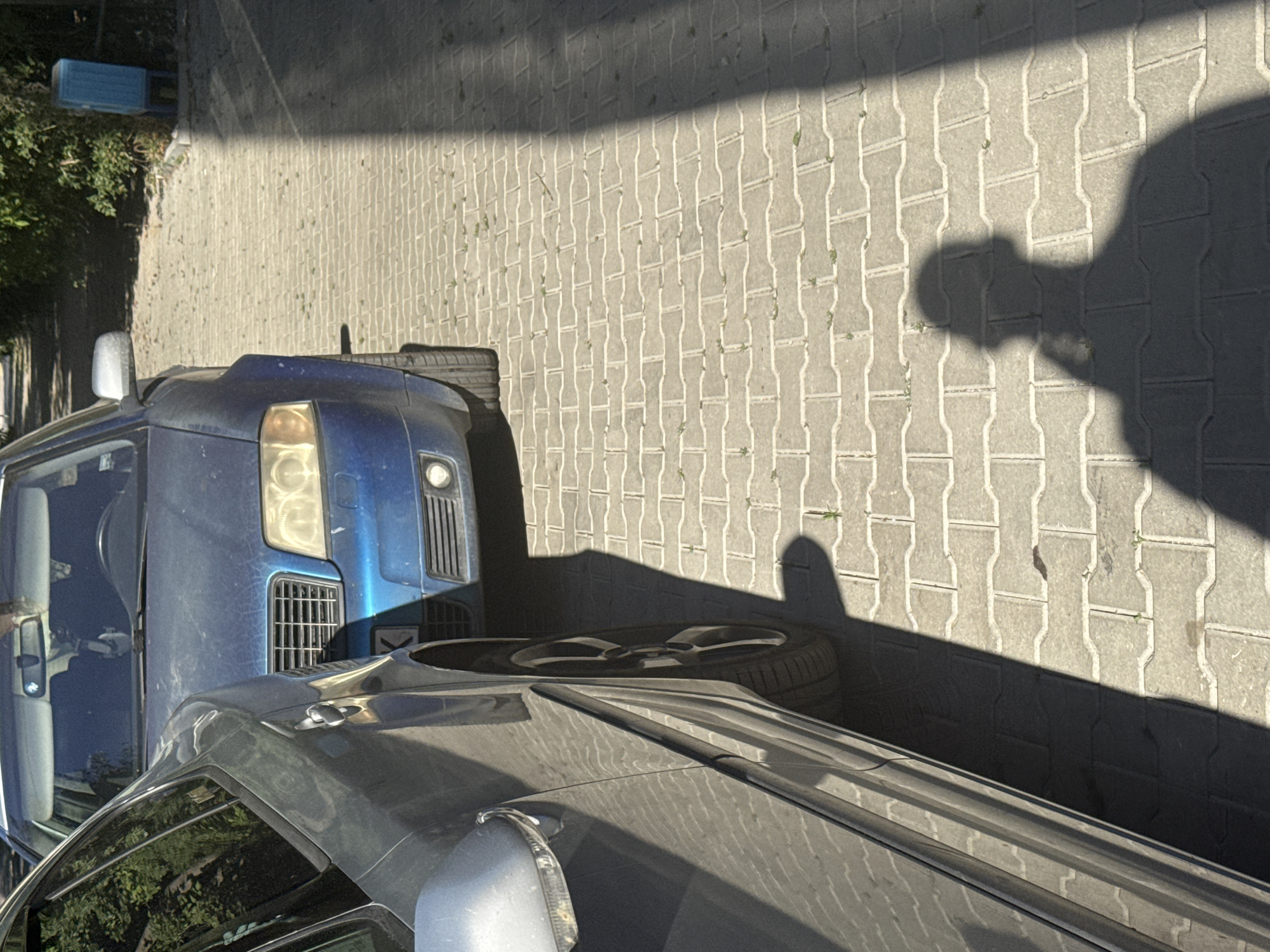 The image shows two cars parked on a pedestrian walkway paved with interlooking grey bricks. A dark blue car is parked in the background, facing forward and slightly angled to the right. A silver car is parked in the foreground, positioned to the left and angled towards the viewer. The silver car's side and wheel are clearly visible. A shadow of a person, holding what appears to be a phone, is cast on the bricks in the foreground, extending from the right side of the frame. The scene is illuminated by sunlight, creating strong shadows. In the upper right corner, a blue trash receptacle is visible amongst green foliage. The environment suggests an outdoor urban setting, likely a residential area or a public walkway in Sofia, Bulgaria.