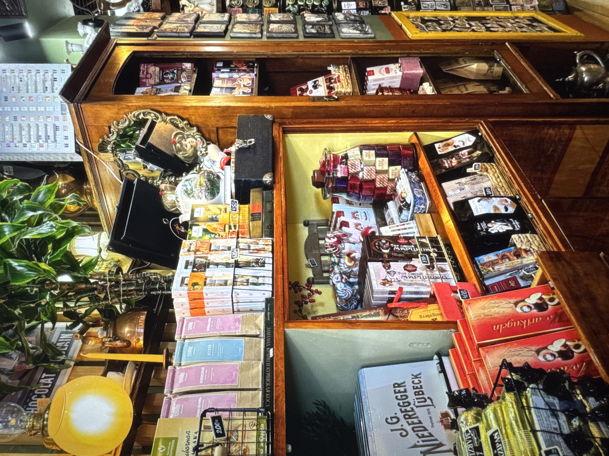 The image depicts a cluttered display of chocolates, teas, and other assorted goods within a shop. The scene is dominated by wooden shelving units and various containers filled with products. In the foreground, shelves are laden with brightly colored boxes of chocolates, bags of tea, and a large glass jar filled with individually wrapped candies. Behind these, a large light blue box with text "J.G. NIEDEREGGER LÜBECK" is visible. The shelves are packed tightly, with some items spilling over or placed precariously.
Further back, a tall wooden cabinet with glass doors showcases more items, including what appear to be packaged chocolates and possibly books or decorative objects. The upper sections of the image reveal a collection of framed pictures or prints mounted on the wall, along with hanging plants and decorative lighting fixtures. A yellow circular lamp with a diffused light source is a prominent feature in the upper left. A kettle and a single candle in a holder are also visible on an upper shelf. The overall impression is one of abundance and a somewhat chaotic, yet curated, retail environment. There are no people visible in the image. The image does not provide any information about the specific location within the city of Novinki, Russia.