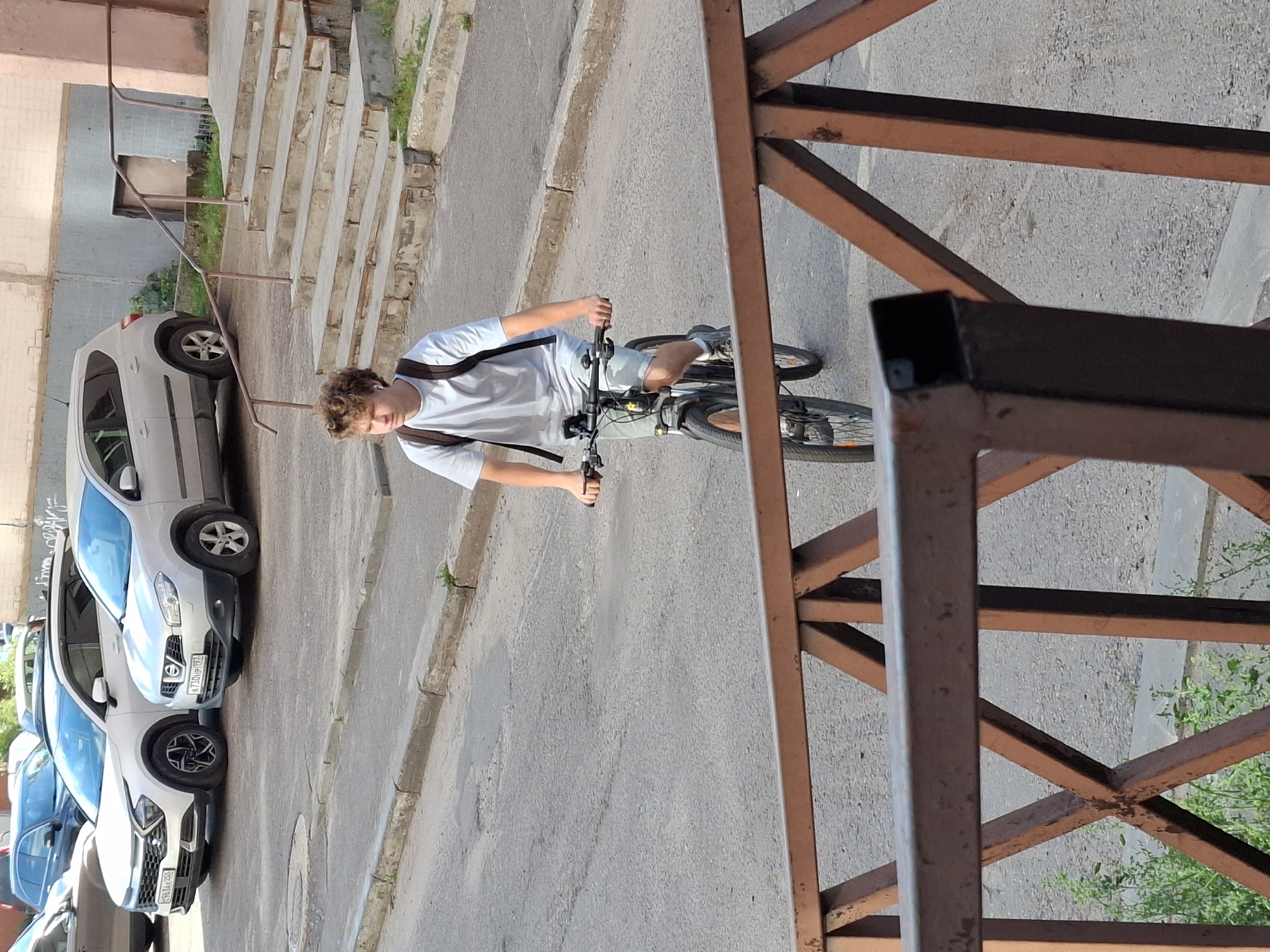 A young male with curly blonde hair rides a bicycle on a paved street. He wears a light grey t-shirt, light blue shorts, and sneakers. He has a dark backpack on his shoulders and is wearing white earbuds. The bicycle has a black frame, black tires, and silver handlebars. In the background, there are two cars parked on the street: a white SUV and a silver hatchback. A building with a light pink facade and concrete stairs with metal railings is visible behind the cars. The scene appears to be in a residential urban area.