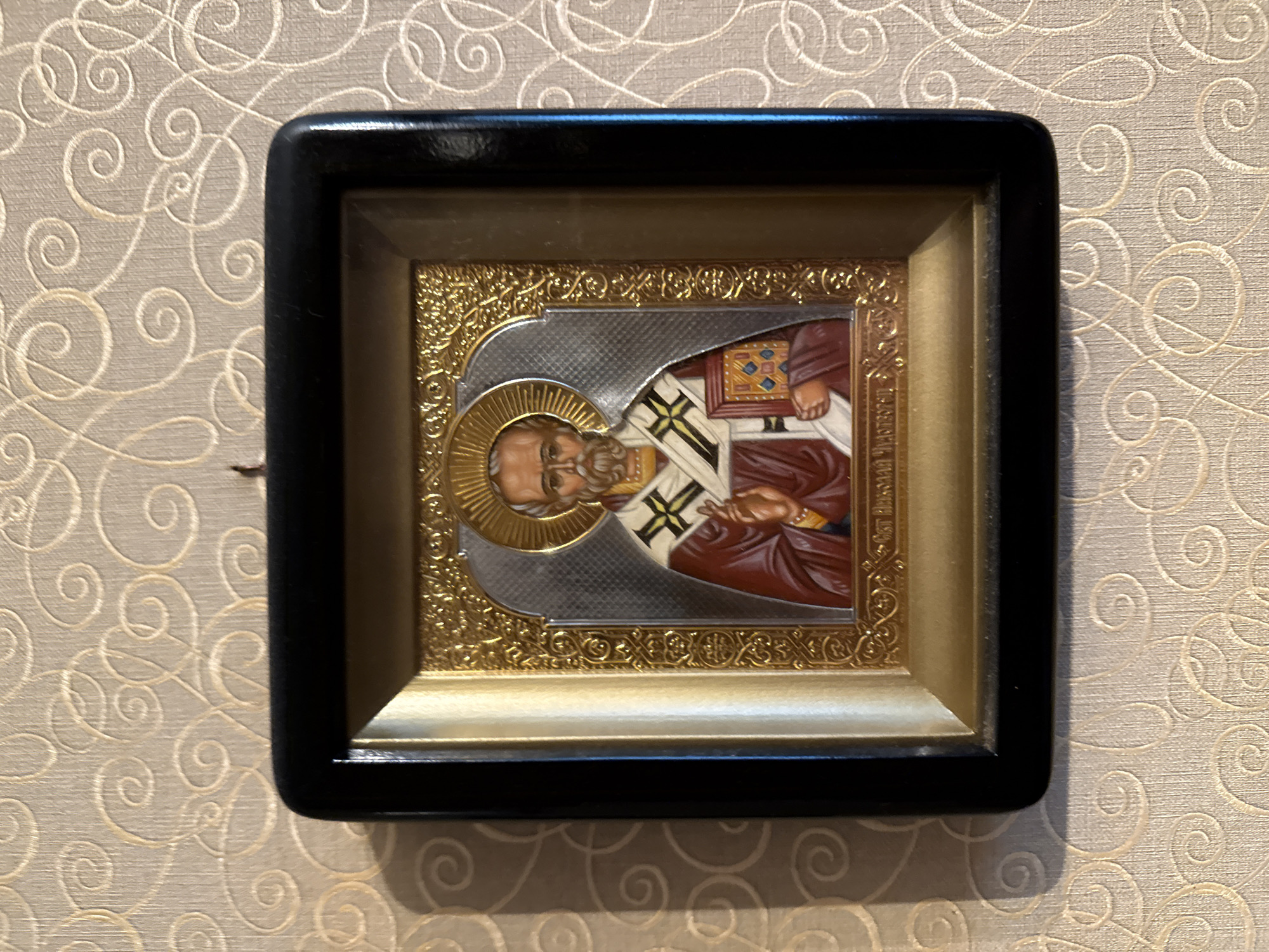 An Orthodox icon of Saint Nicholas is framed in black and gold and hangs on a wall with a beige, swirled wallpaper pattern. The icon depicts Saint Nicholas wearing a red robe and a white and gold stole, holding a book in his left hand and raising his right hand in blessing. He has a halo behind his head. The icon is centrally located in the frame, which is mounted on a textured wallpaper with a repeating curvilinear pattern. The overall scene is a close-up view of the icon on the wall. This image was taken in the city of Rudamina, Lithuania.