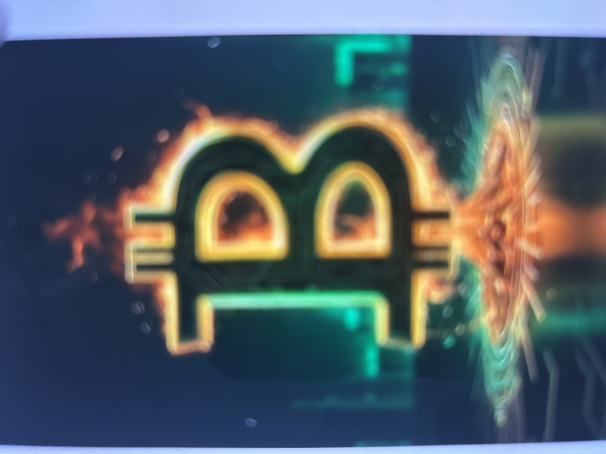 The image is a vertical, close-up shot of a Bitcoin symbol engulfed in flames. The symbol itself is rendered in a neon-like style, with green and yellow hues, and it appears to be three-dimensional. The flames are depicted with vibrant reds and oranges, surrounding the top and bottom of the symbol. The background is dark and out of focus, with hints of neon green lines and geometric shapes, suggesting a digital or futuristic cityscape. There are also some out-of-focus bright spots, possibly representing lights or reflections. The image does not contain any identifiable people, objects, or specific environmental details that would allow for the identification of a location in Gelendzhik, Russia.