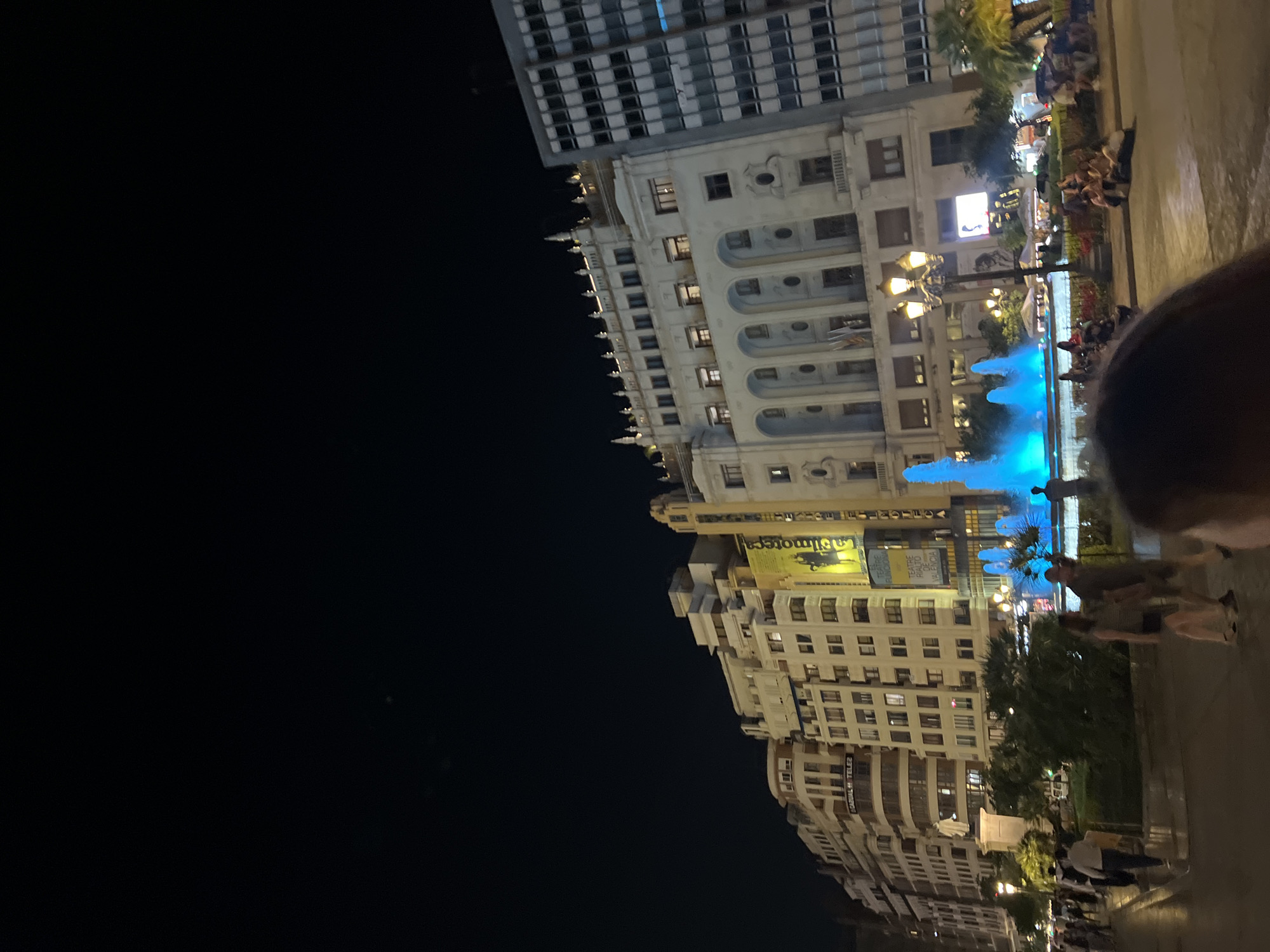 The image depicts a nighttime scene in Valencia, Spain, centered around a fountain with blue lights in the Plaza del Ayuntamiento. The plaza is surrounded by buildings, including the Ayuntamiento (City Hall) and other ornate structures. People are walking and sitting around the fountain. There are streetlights illuminating the area.