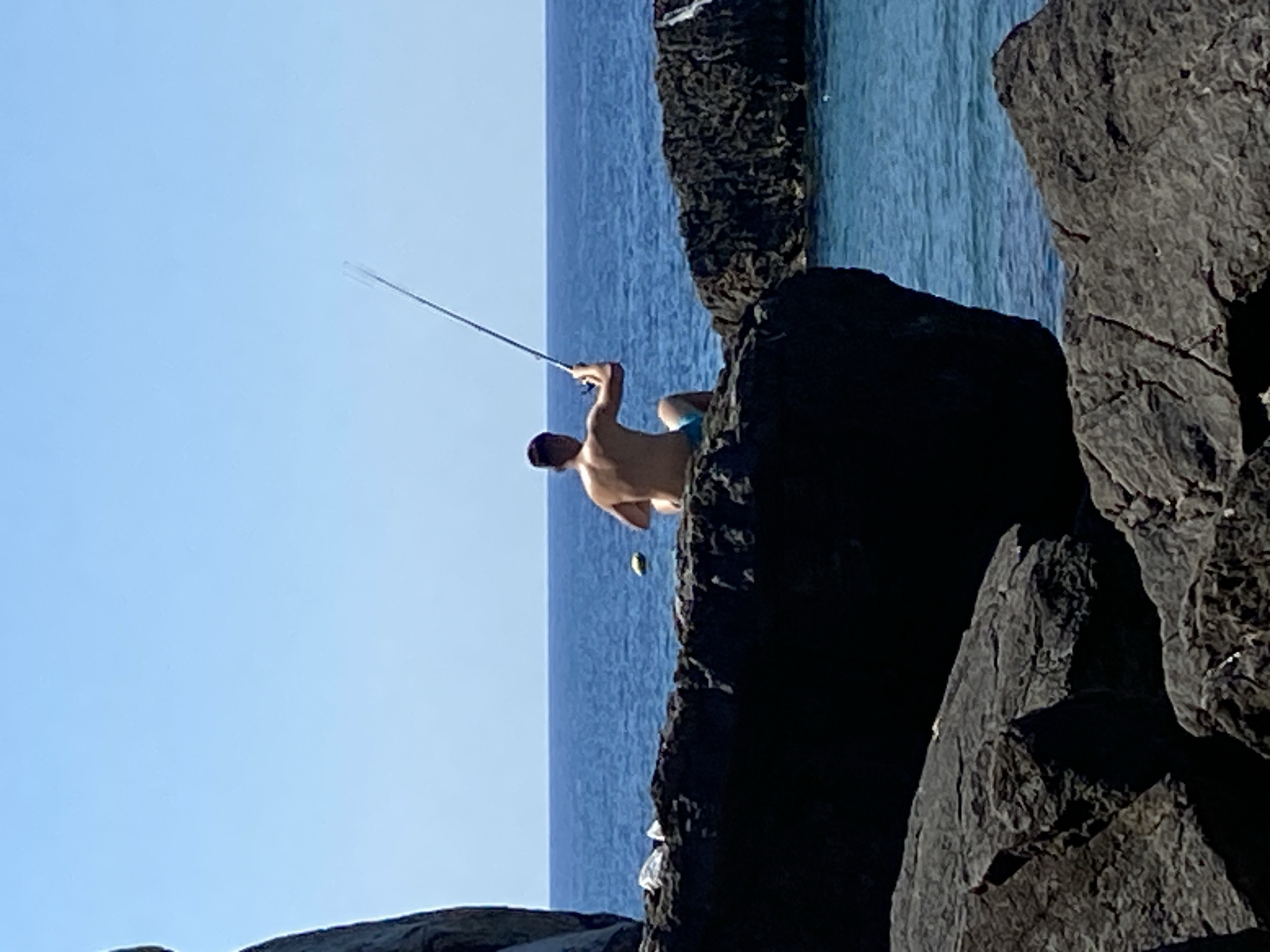 A shirtless person wearing blue swim trunks sits on dark rocks by the sea, holding a fishing rod. The person is facing away from the camera and looking out at the water. The ocean is a deep blue, and the sky is a clear, bright blue. In the foreground, large, dark rocks dominate the scene. There is a yellow object floating in the water near the person. The overall scene depicts a sunny day spent fishing by the coast, possibly in Budva, Montenegro, given the typical rocky coastlines and clear blue waters found in that region.