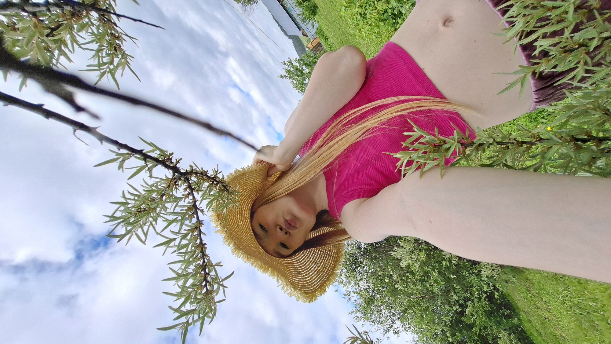 A young woman with long blonde hair, wearing a bright pink crop top and a straw hat, is taking a selfie outdoors. She is outdoors, possibly in the village of Bolshoy Bukor, Russia, as indicated by a distant house and structures visible in the background, nestled amongst trees and green foliage. The woman is positioned in the foreground, with a branch of a tree, possibly sea buckthorn, with small green leaves, partially obscuring her face. Her arm is extended, holding the camera for the selfie, and her lips are pursed. The sky above is a mix of blue with fluffy white clouds. The overall impression is a casual, outdoor portrait.
