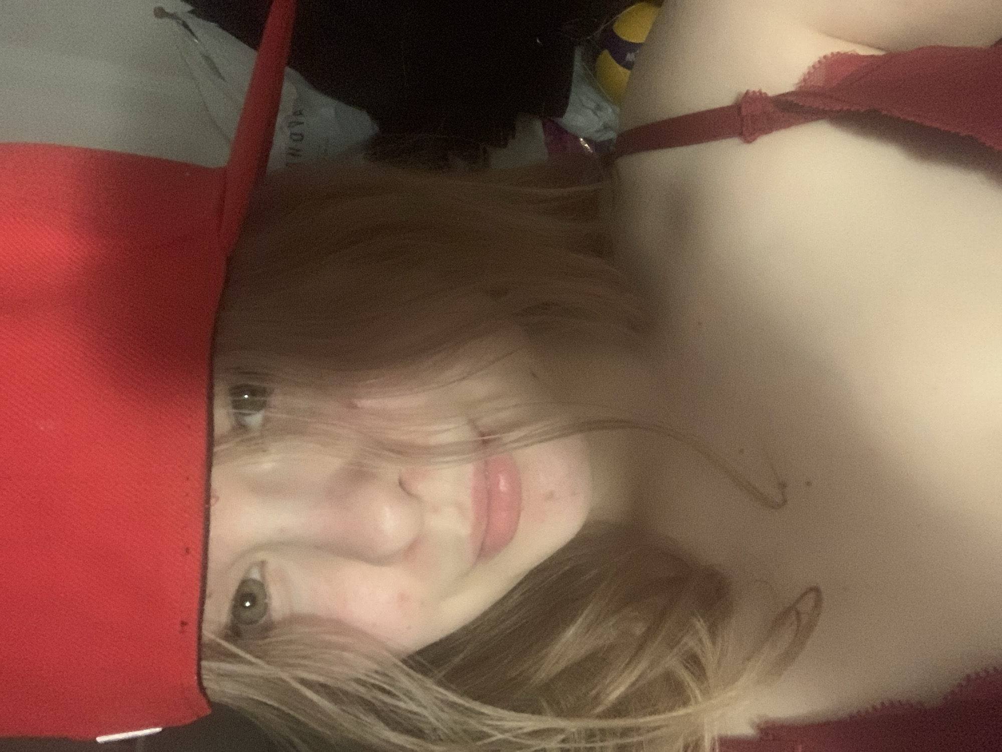 This medium-shot, portrait-oriented image, taken in Saint Petersburg, Russia, features a young person lying down and looking directly at the camera, likely capturing a selfie. The subject, appearing to be female, has light brown or blonde hair, fair skin, and green or hazel eyes. They are wearing a prominent red baseball cap, which covers the left side of their head and part of their forehead. A red or burgundy bra is visible on their upper body.
The person's expression is subtle, with a slight, almost imperceptible smile and softly pink lips. Their hair is somewhat disheveled, falling around their face. The background is dimly lit and appears to be an indoor setting, possibly a bedroom, characterized by dark, indistinct shapes and various cluttered items. Hints of other objects are visible, including a light-colored item with some partially obscured and unreadable text (possibly "ADNA" or "NOVA") and a yellow and purple object, potentially a ball, further back in the frame. The overall lighting is low, creating a casual and slightly intimate atmosphere. The image itself is rotated clockwise, positioning the subject's head on the left side of the frame.