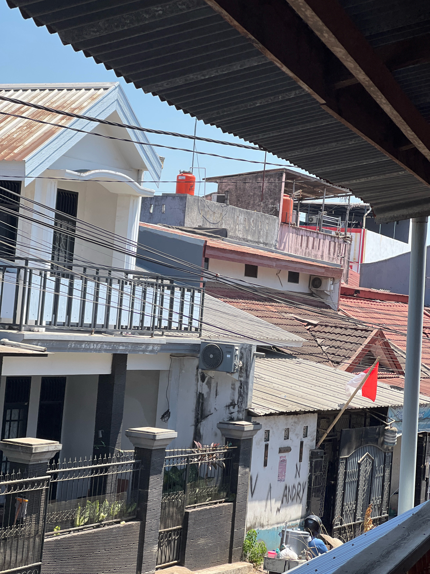 This is a photograph of a residential area in Makassar, Indonesia. The scene is densely packed with houses, characterized by corrugated metal roofs and a mix of building materials. In the foreground, a black metal fence encloses a small garden area with some potted plants. Two stone pillars flank the fence.
Behind the fence, the lower level of a white house is visible, featuring dark-framed windows and a balcony with a similar black metal railing. Above this, several other houses are stacked and clustered, with varying roof heights and colors, predominantly reddish-brown tiles and weathered metal. Electrical wires are strung haphazardly between buildings.
An orange water tank is prominent on top of one of the buildings. A small Indonesian flag, red and white, is mounted on a bamboo pole and flies from a house in the middle ground. The walls of some houses show signs of wear and graffiti, including the name "ANDRY" scrawled in black spray paint. An air conditioning unit is mounted on one of the walls. In the lower right portion of the image, part of a blue scooter or motorcycle is visible. The overall impression is a bustling, urban residential environment typical of Makassar.