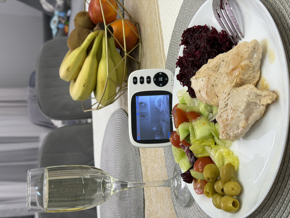 The scene captures a dining table in an indoor environment, located in Zelenaya Polyana, Russia. A white rectangular plate in the foreground contains a meal consisting of two pieces of cooked chicken breast, a portion of shredded red beetroot, a mixed green salad with lettuce, cucumber slices, and cherry tomatoes, and a cluster of green olives. A silver fork rests on the plate next to the beetroot. To the left of the plate, a clear champagne flute holds a light-colored, effervescent beverage. Centrally positioned on the table, a white baby monitor with a black screen displays a low-light image of an infant sleeping in a crib within a room, with the time "19:35" visible on the screen. The monitor features white control buttons and indicator lights on its right side. Behind the monitor, a gold-colored, geometric wire fruit basket holds a cluster of yellow bananas, two brown kiwis, and three orange-colored fruits. The table is covered with a light-colored tablecloth, and a round, woven gray placemat is visible under the champagne flute. The background is blurred, showing grey upholstered dining chairs and light-colored curtains.