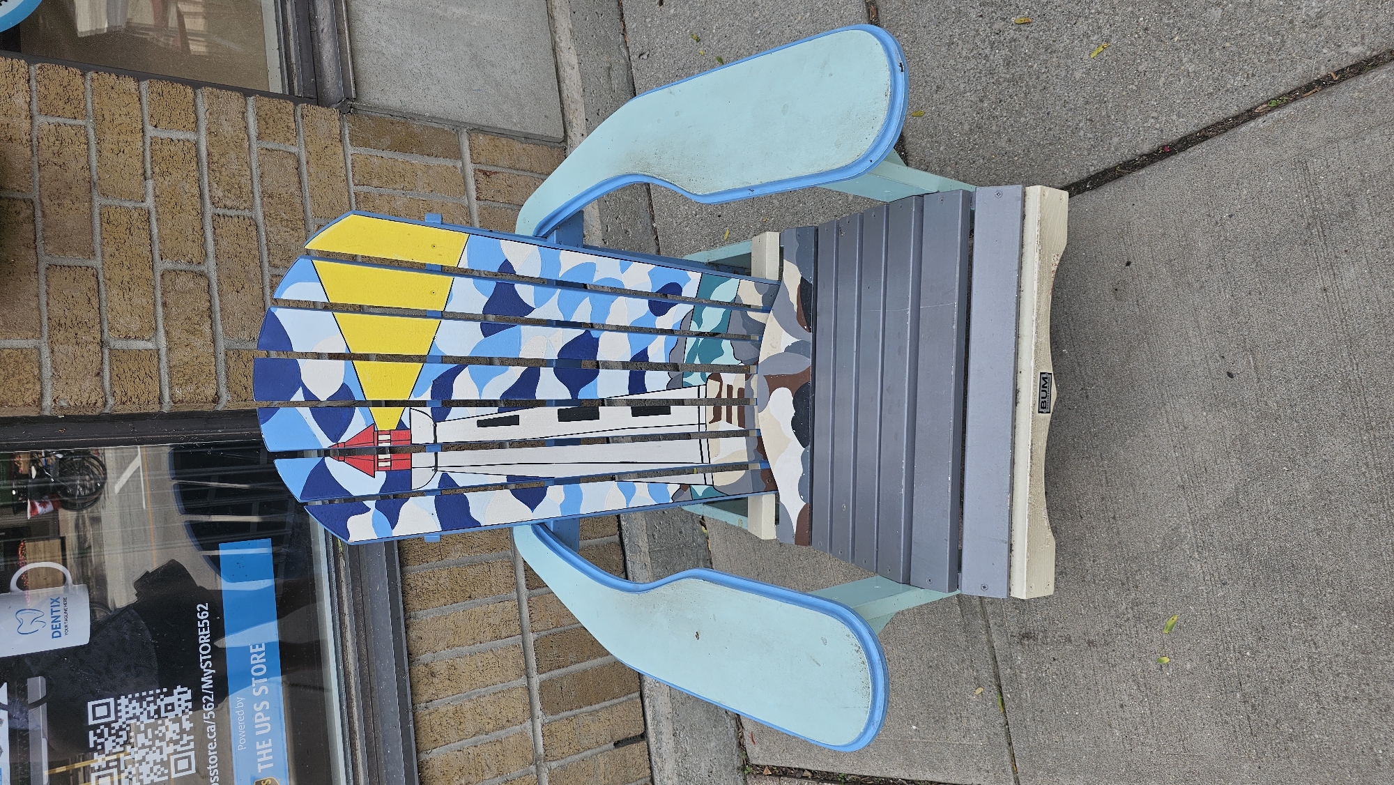This media file shows a top-down view of a decorated Adirondack chair placed on a concrete sidewalk outside a building. The chair's seat and back are painted with a stylized design featuring a lighthouse, a yellow sun, and abstract blue and white shapes, possibly representing water or a sky. The armrests are painted a light blue. The back of the chair has a ribbed, gray panel. A small black label with the word "BUM" is visible on the side of the chair.
The background includes a brick wall and a storefront window. Reflected in the window are a white mug with a blue logo that says "DENTIX YOUR HEALTHIER SMILE," a QR code, and text indicating "Powered by: store.ca/562/MySTORE562" and "THE UPS STORE." This suggests the chair is located outside a UPS Store.
The lighting appears to be natural, suggesting it is daytime. No people are visible in the frame. The scene is a common urban or commercial outdoor setting. The chair is the primary subject, presented as a piece of art or public installation. The overall impression is one of a publicly accessible, perhaps decorative, object in a commercial area.