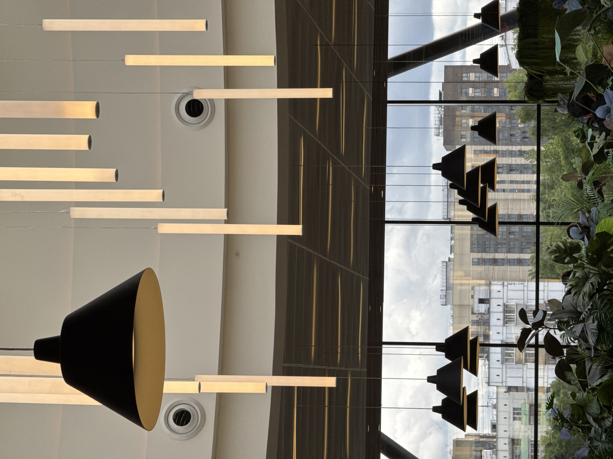 A low-angle, indoor shot showcases a modern interior juxtaposed against a cityscape. The foreground features an array of horizontal tube lights and one downward-facing, cone-shaped lamp with a black exterior and gold interior. The ceiling is white with circular fixtures that may be vents or lights. Through large, multi-paned windows, a cityscape is visible, including several apartment buildings and office blocks. Hanging lamps, similar to the foreground lamp, are silhouetted against the urban backdrop. Lush greenery is positioned near the window. The location is in Moscow, Russia.