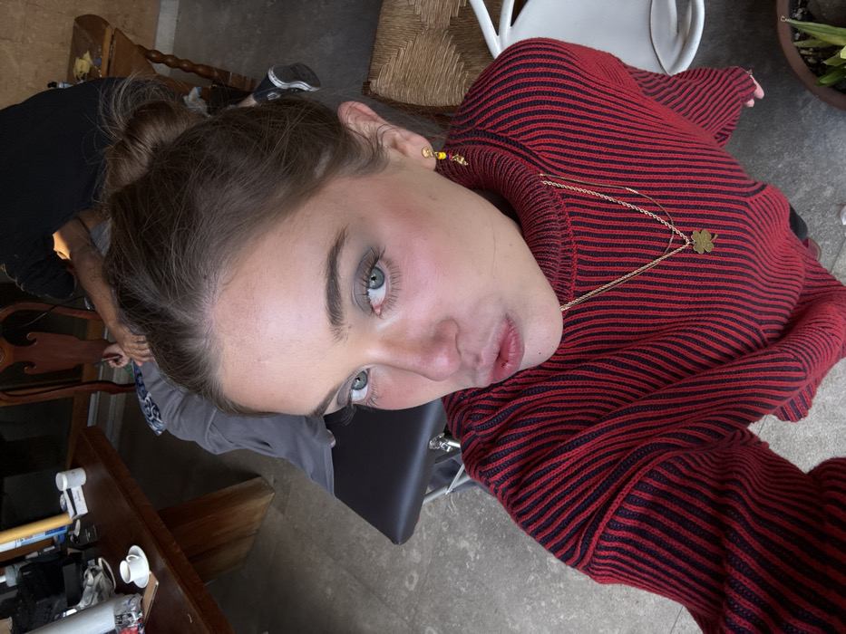 A woman looks up at the camera, her head tilted slightly. She has fair skin, light brown hair in a bun, and blue eyes. She wears a red and black striped turtleneck sweater and a gold necklace with a clover pendant. A small gold earring is visible in her left ear. Various objects are scattered around, including furniture, a plant pot, and cleaning supplies. The floor appears to be concrete.