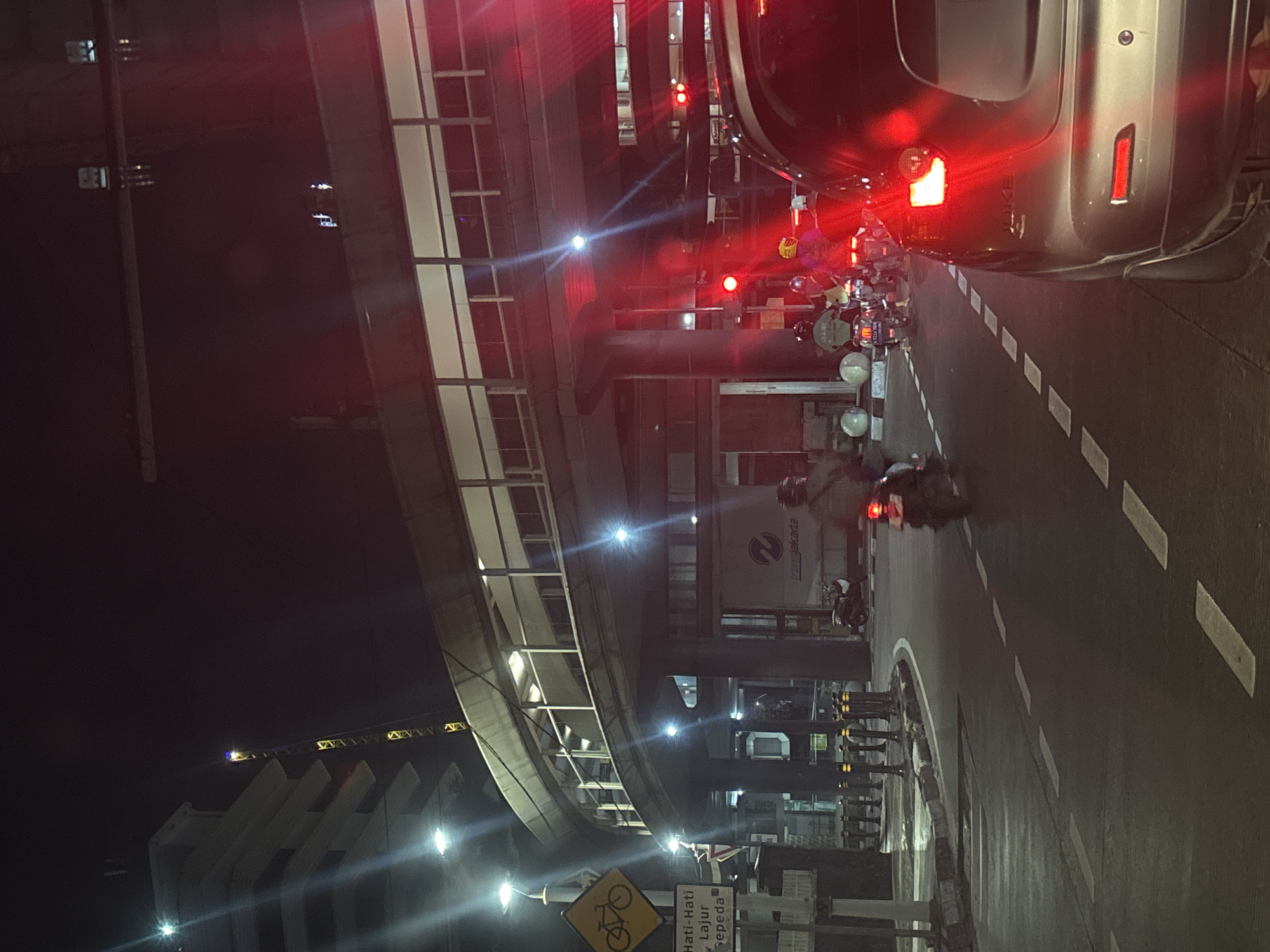 The image, rotated ninety degrees clockwise, shows a night scene in South Jakarta, Indonesia. Cars are stopped at a red light, and motorcycles weave through the traffic. An elevated pedestrian walkway curves overhead, illuminated by streetlights. A sign indicates a bicycle lane. The street is marked with dashed lines. A silver car is partially visible in the right foreground. Construction appears to be ongoing, with a crane silhouetted against the dark sky. A sign with “Transjakarta” is visible near the walkway.