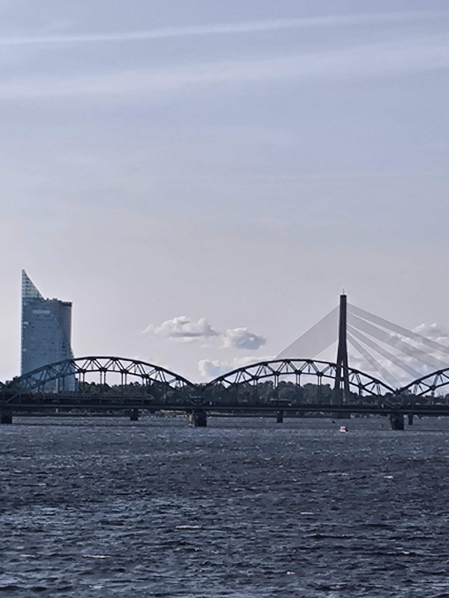 The image depicts a wide shot of the Daugava River in Riga, Latvia, under a cloudy sky. On the left, a modern, sharp-angled skyscraper with a bluish-green facade stands prominently. In the middle ground, a dark metal arched bridge spans the river, its intricate truss structure visible. To the right of this bridge, a more modern cable-stayed bridge with a tall, single pylon is partially visible. The water of the river is dark and choppy, with subtle ripples and a few faint reflections. The background is dominated by a pale sky with scattered, wispy clouds. No people are visible in the frame.