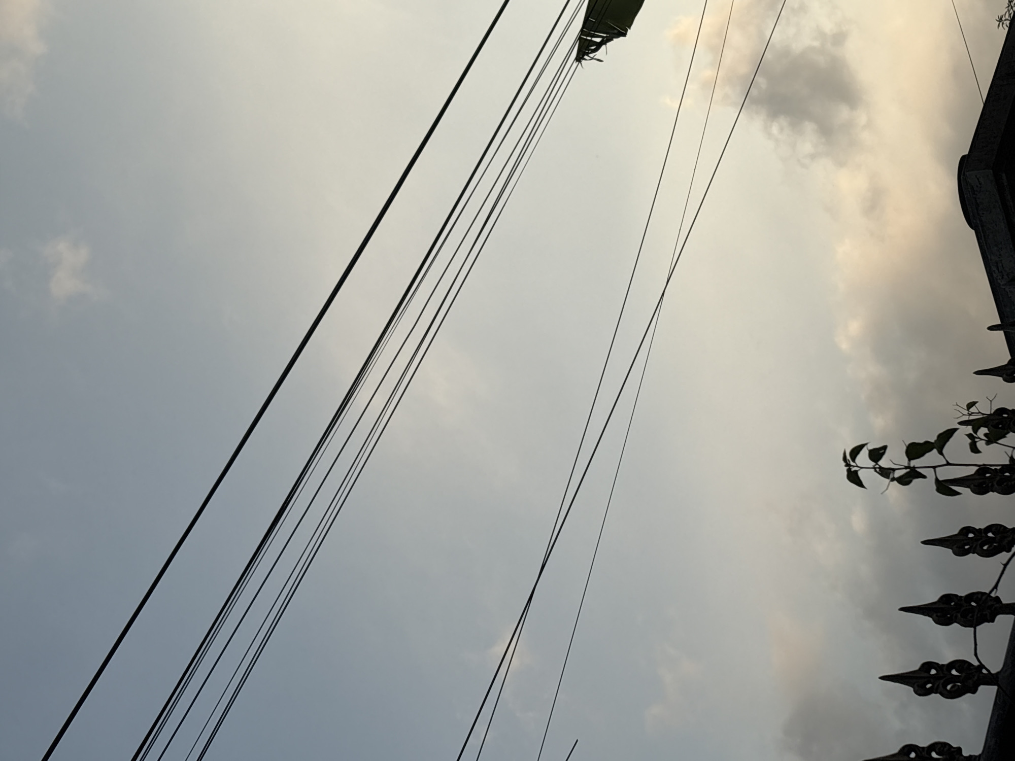 The image depicts a clear sky with scattered clouds, with multiple power lines diagonally crossing the frame. In the lower portion of the image, a dark, decorative fence with pointed finials is visible, along with a small, silhouetted plant. The scene appears to be a street view, possibly in Palimanan, Indonesia, given the context. The lighting suggests it is either dawn or dusk. There are no people or animals visible in the image.