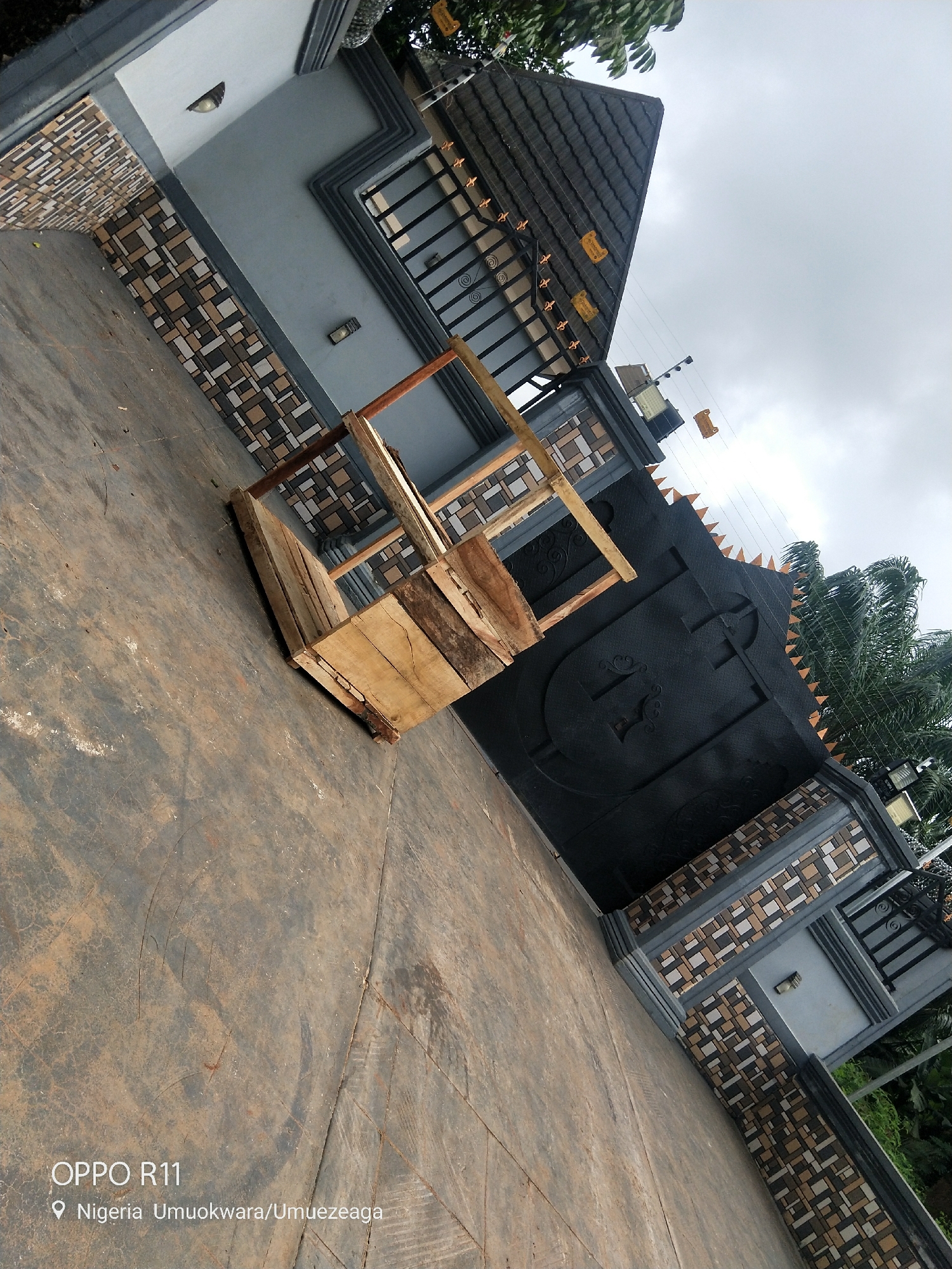 A wooden structure, possibly a bench or a makeshift seating, made of rough planks is placed on a paved ground. The structure is in the foreground, tilted to the left. Behind it is a large, ornate black gate with decorative patterns and what appears to be a cross emblem. The gate is flanked by patterned walls and a tiled fence. The overall scene is outdoors, in what looks like a residential or commercial property entrance. The sky is overcast, suggesting it might be a cloudy day. There are trees visible in the background, to the right of the gate. The lighting suggests it's daytime. Visible text in the lower left corner reads "OPPO R11" and "Nigeria Umuokwara/Umuezeaga". The setting appears to be Akokwa, Nigeria. No people are visible in the image. The scene is static, with no particular activity taking place. The focus is on the architectural elements and the wooden object.